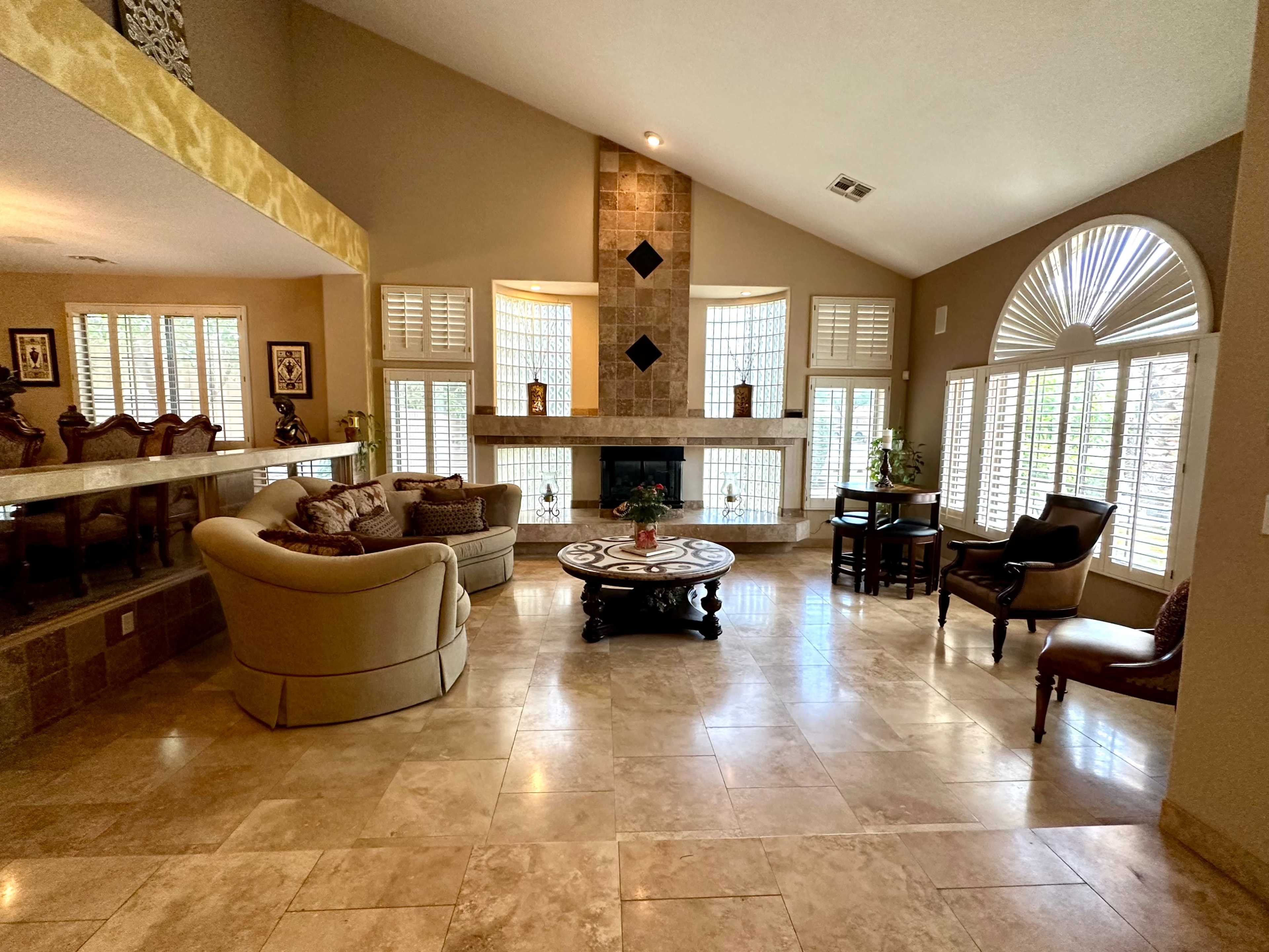 The image shows a spacious living area with a stone fireplace, large windows, and a combination of seating arrangements including a sofa, chairs, and a coffee table.