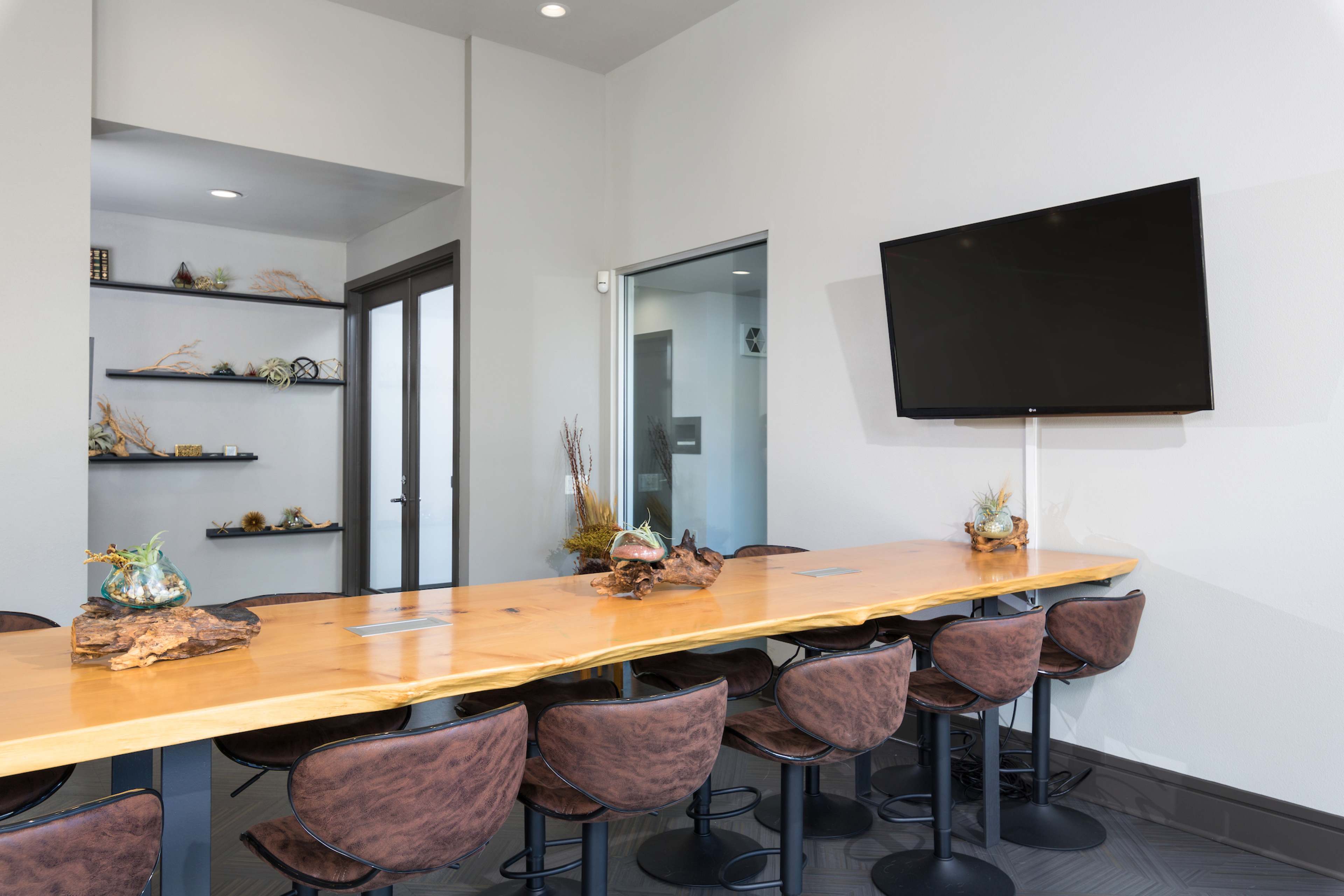 A modern meeting room features a long wooden table with bar stools, a television mounted on the wall, and decorative shelves.