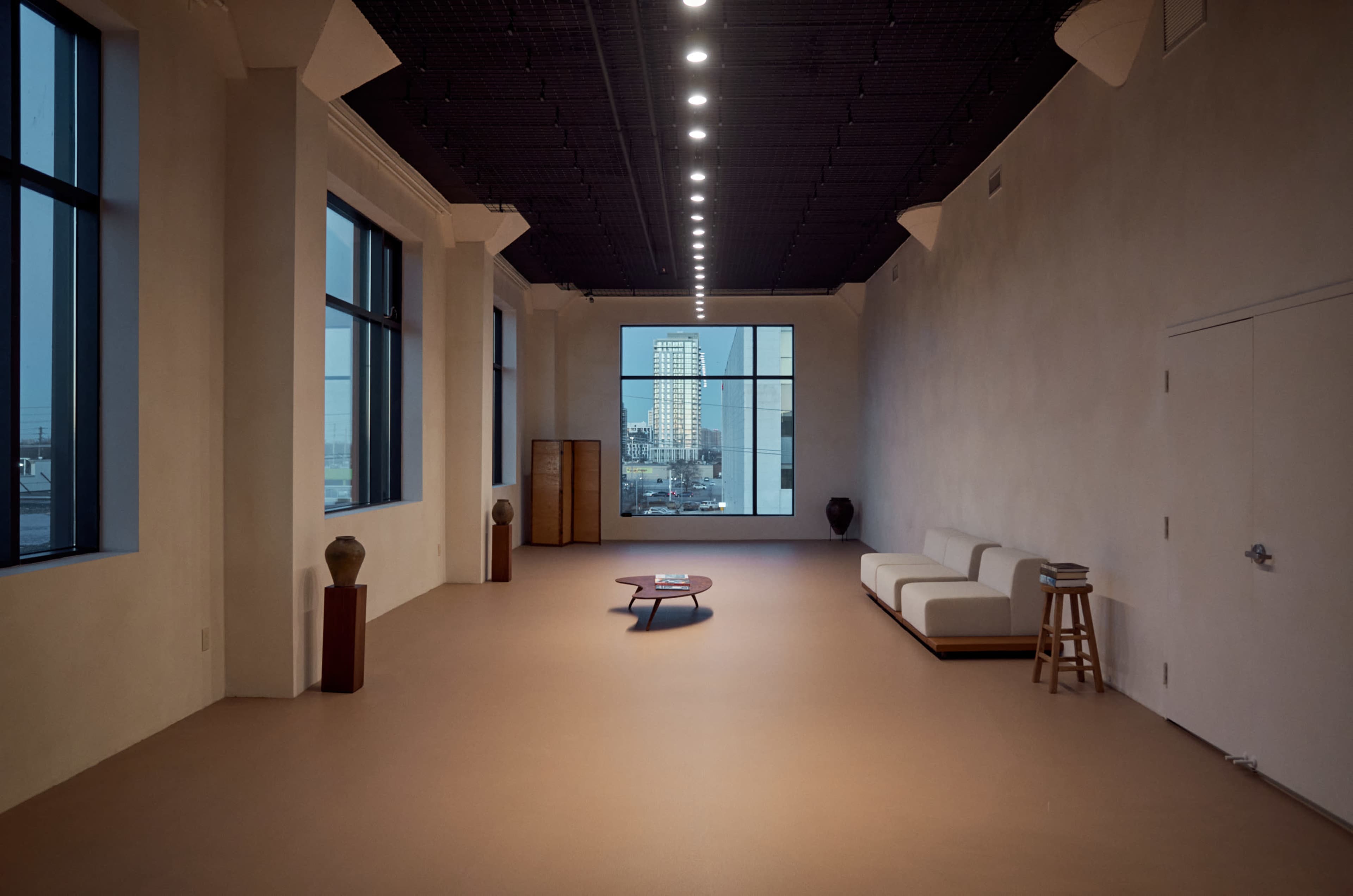 The image depicts a spacious, minimalist interior with large windows and a view of a city skyline, featuring a simple sofa, a low table, and a few decorative elements.