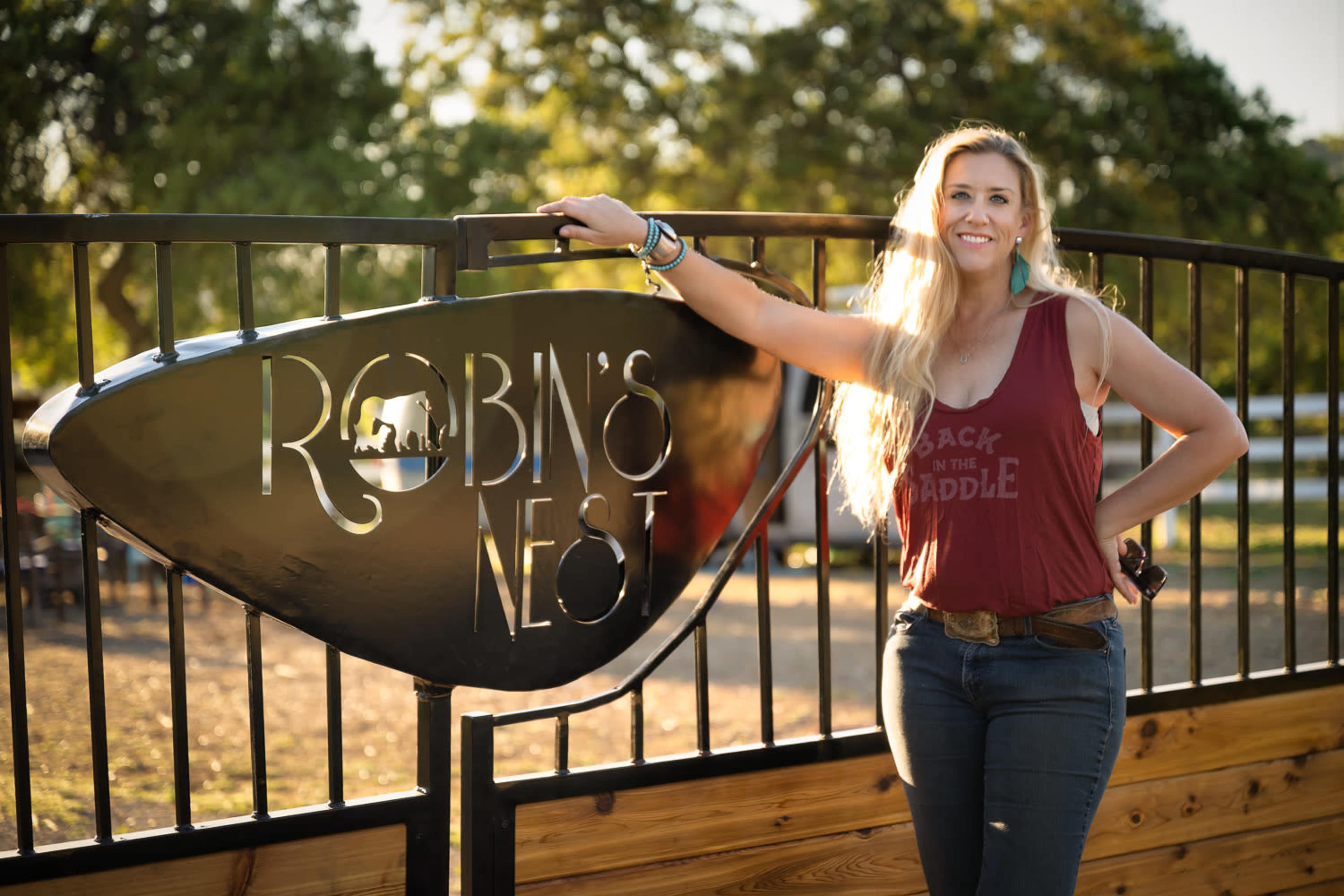 A woman stands beside a modern metal sign that reads "Robin's Nest," with an outdoor setting in the background.