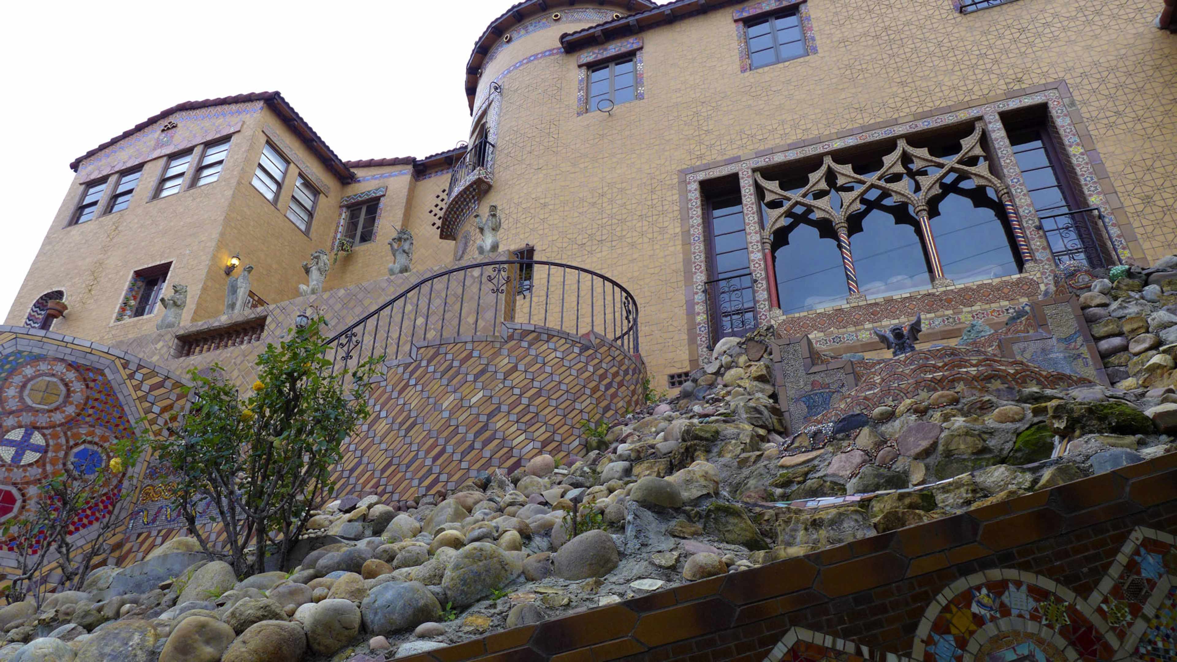 A multi-level building with a stone facade, decorative mosaics, and a stairway surrounded by a landscaped area of rocks and plants.