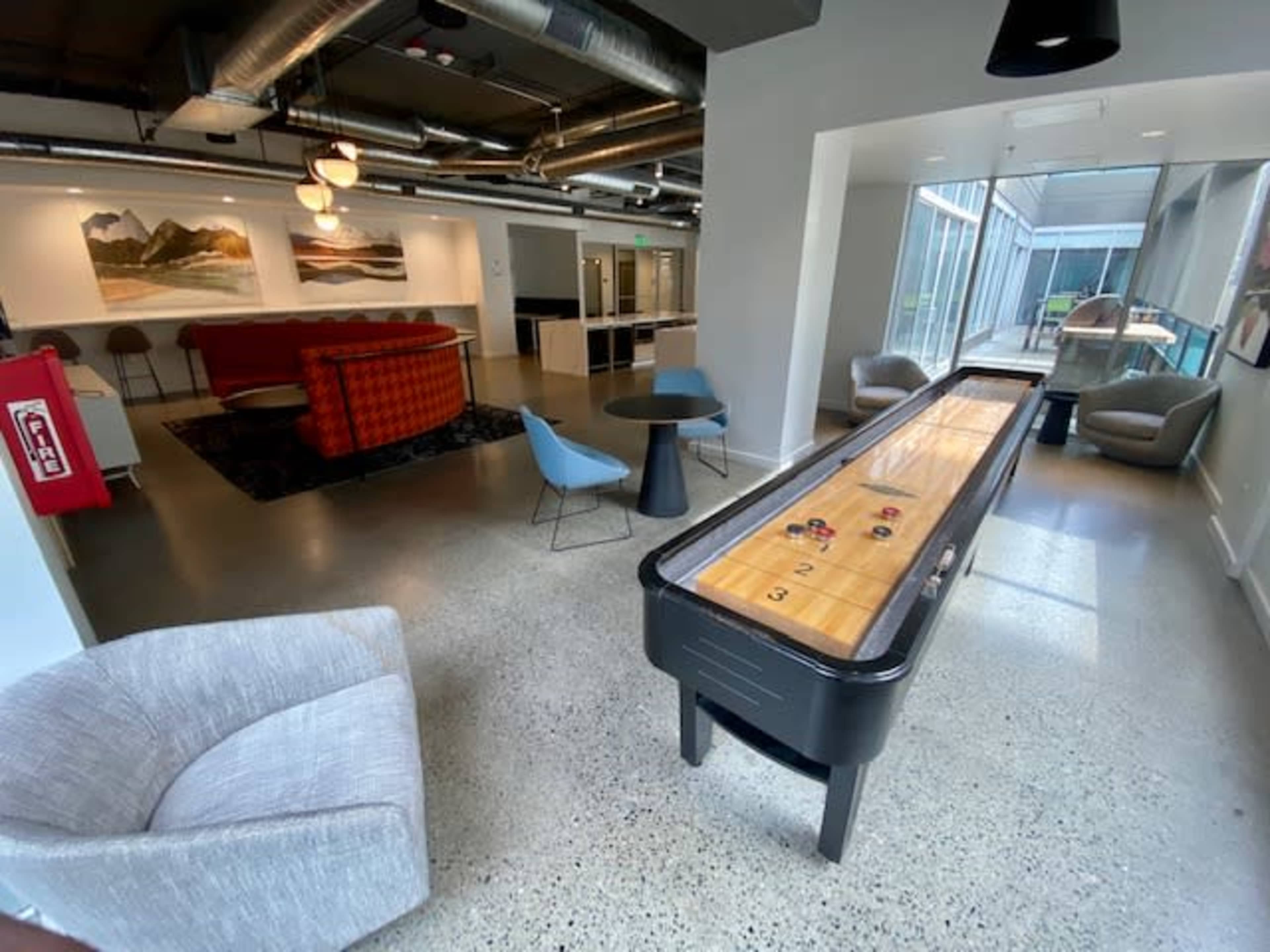 A modern lounge area with a shuffleboard table, comfortable seating, and large windows letting in natural light.