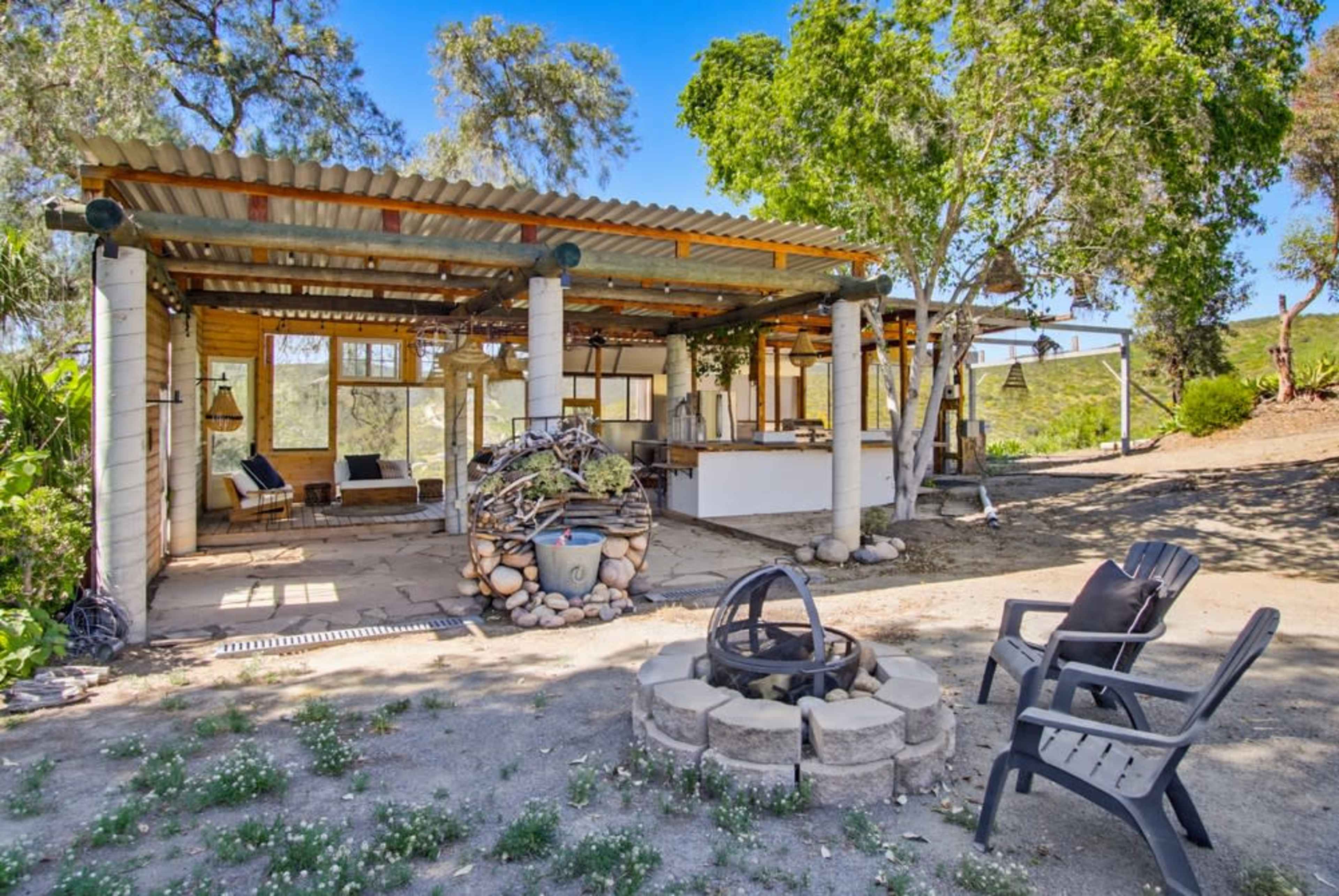 The image shows a rustic outdoor living space with a stone fire pit, seating area, and a partially covered structure surrounded by trees.