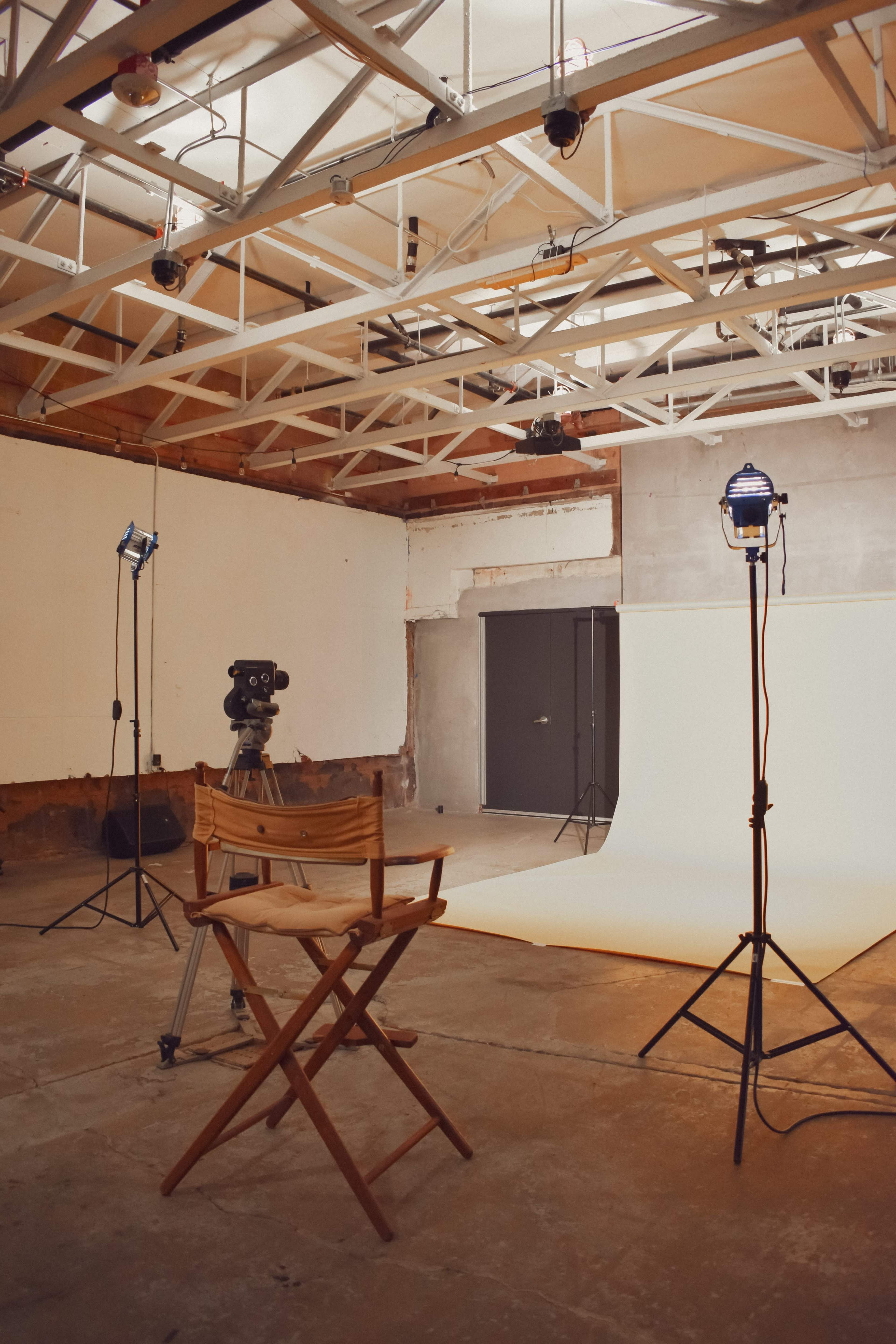 A filmmaking studio features a wooden director's chair facing a plain backdrop, flanked by lighting equipment and a vintage film camera on a tripod.