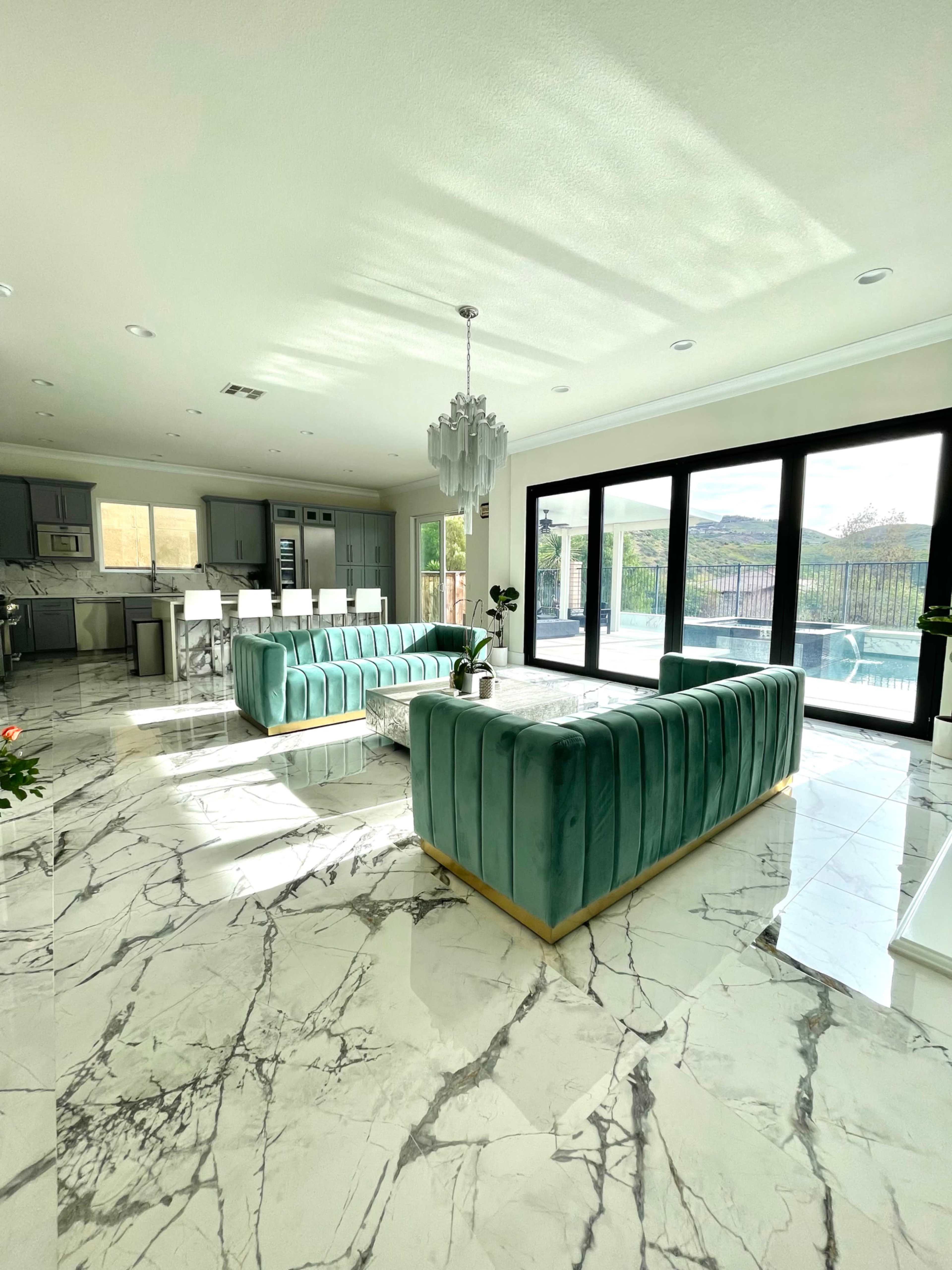 A modern living room with green velvet sofas arranged around a marble coffee table, large windows showcasing a view of the outdoor pool area, and a sleek kitchen in the background.