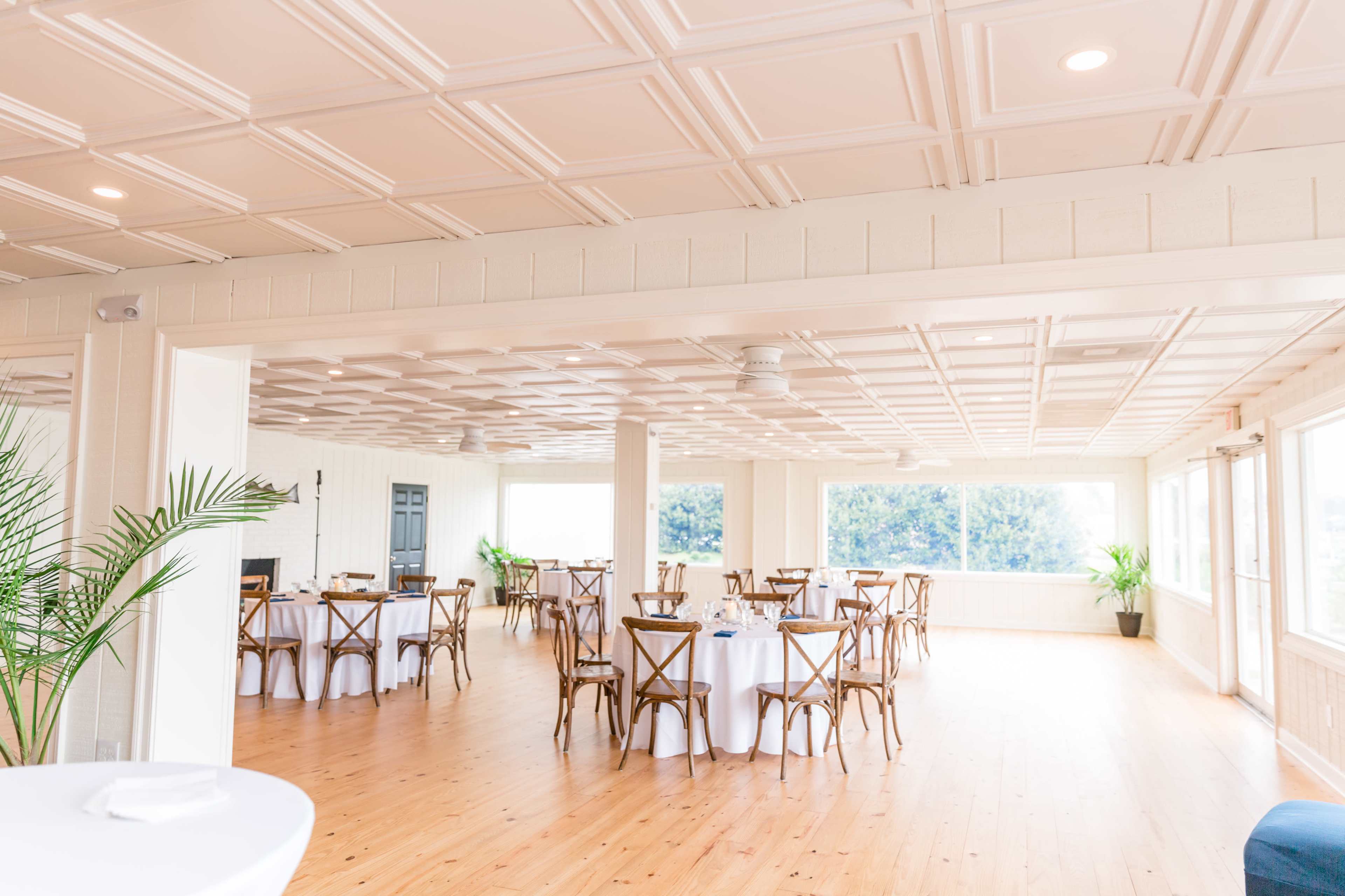 A spacious dining area features wooden floors, round tables with white tablecloths, and large windows allowing natural light.