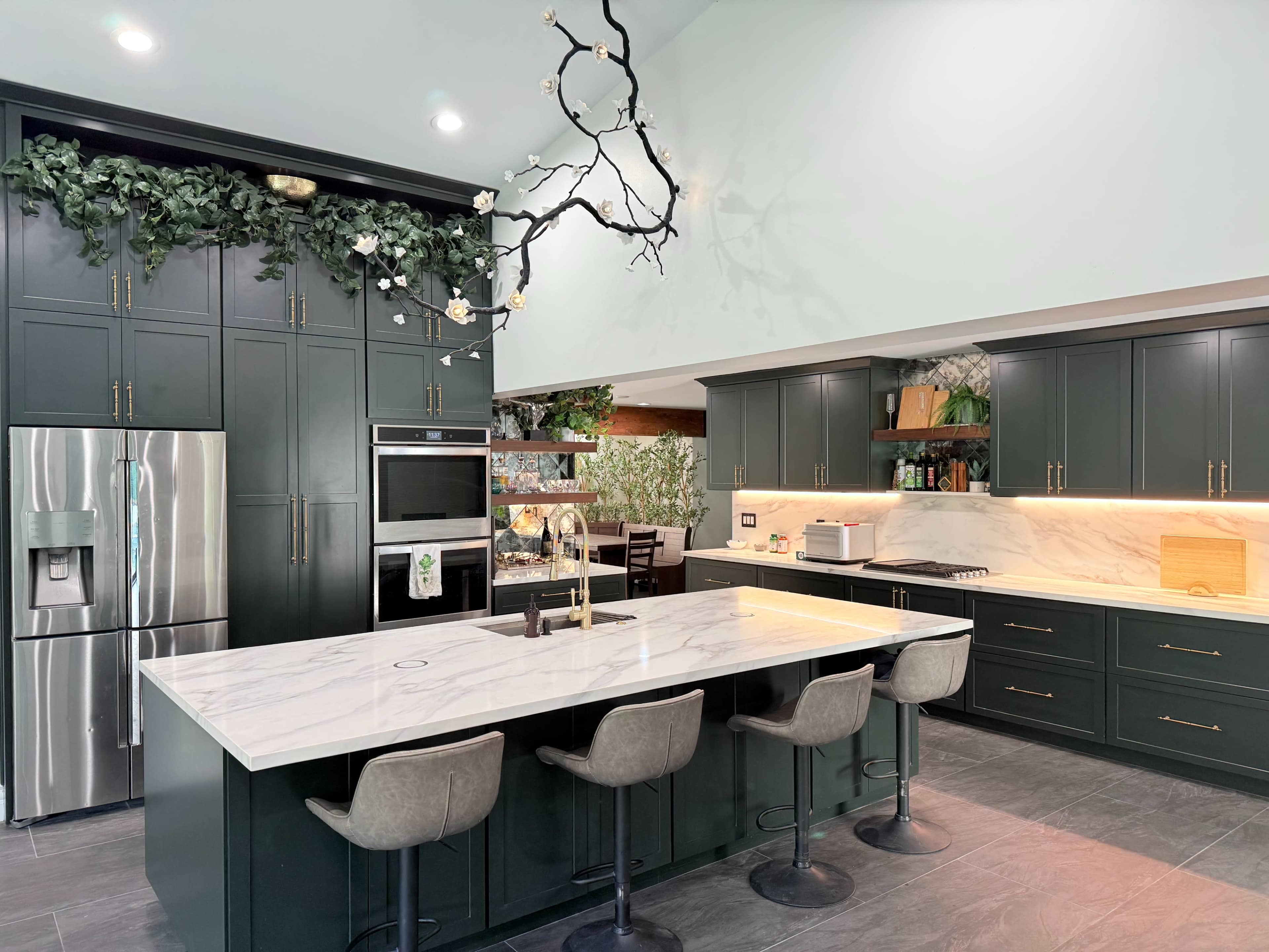 The kitchen features dark green cabinetry, a large marble island with seating, stainless steel appliances, and a decorative branch fixture overhead.