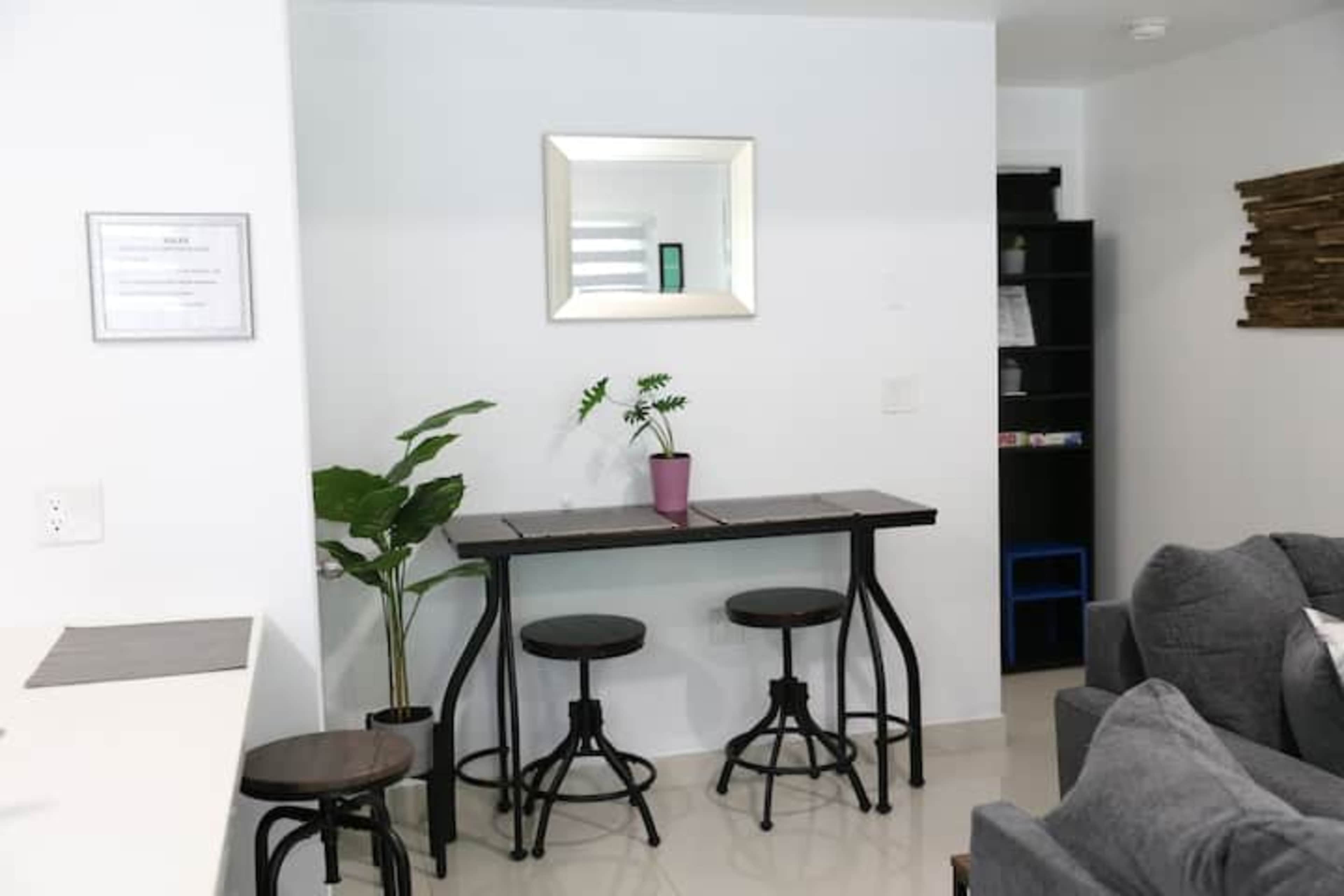 A small dining area features a black table with two stools, a potted plant, and a mirror on the wall.