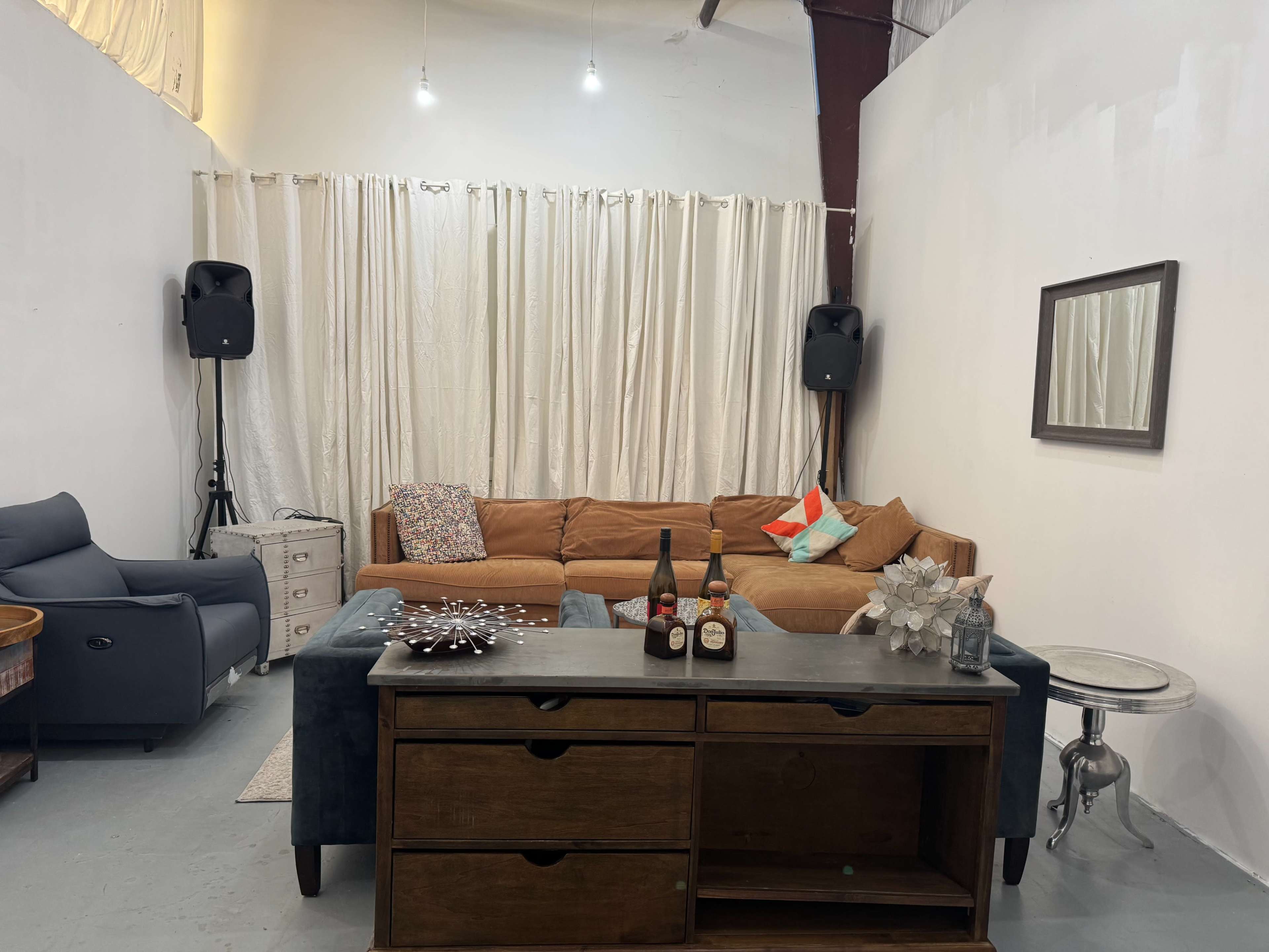 The image shows a cozy living room with a brown sectional couch, a wooden side table, and decorative elements like a mirror and bottles on the table, all set against a white wall and curtains.