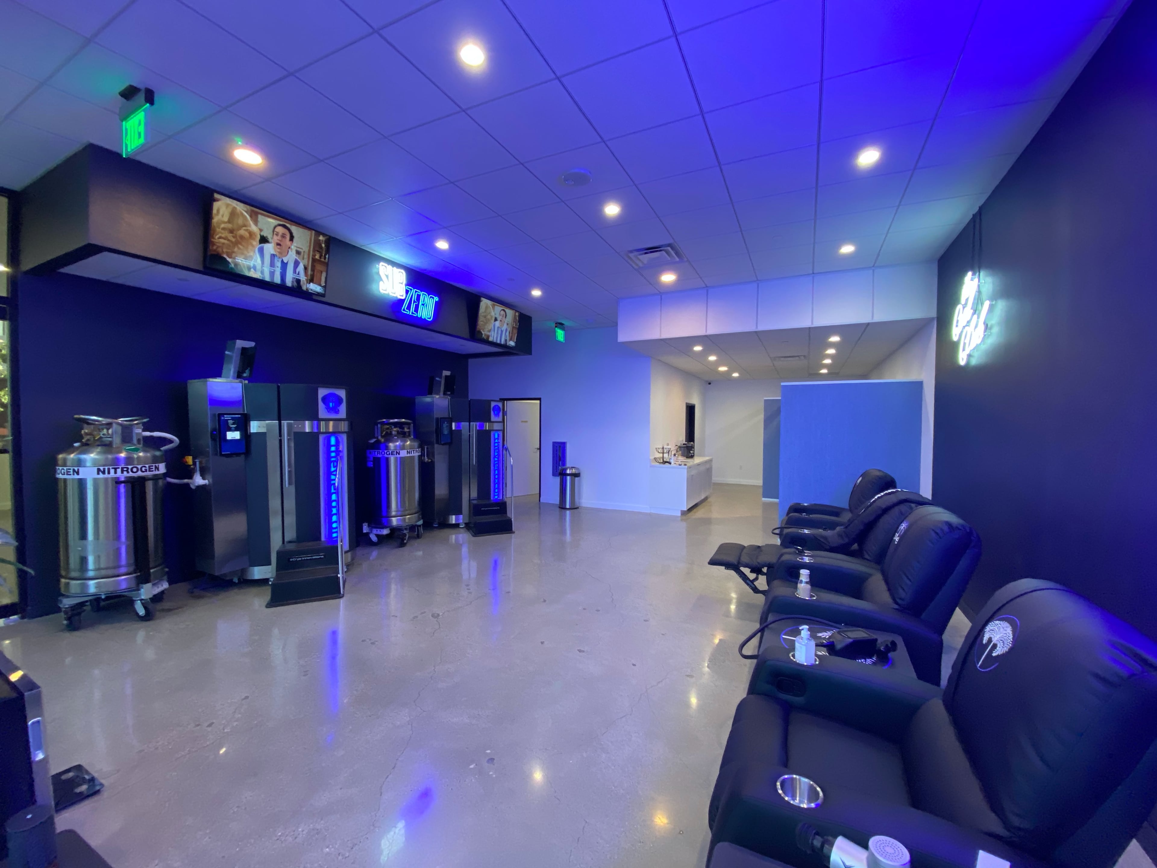 The image shows a modern wellness center with sleek black chairs, nitrogen tanks, and large screens displaying information on the walls.