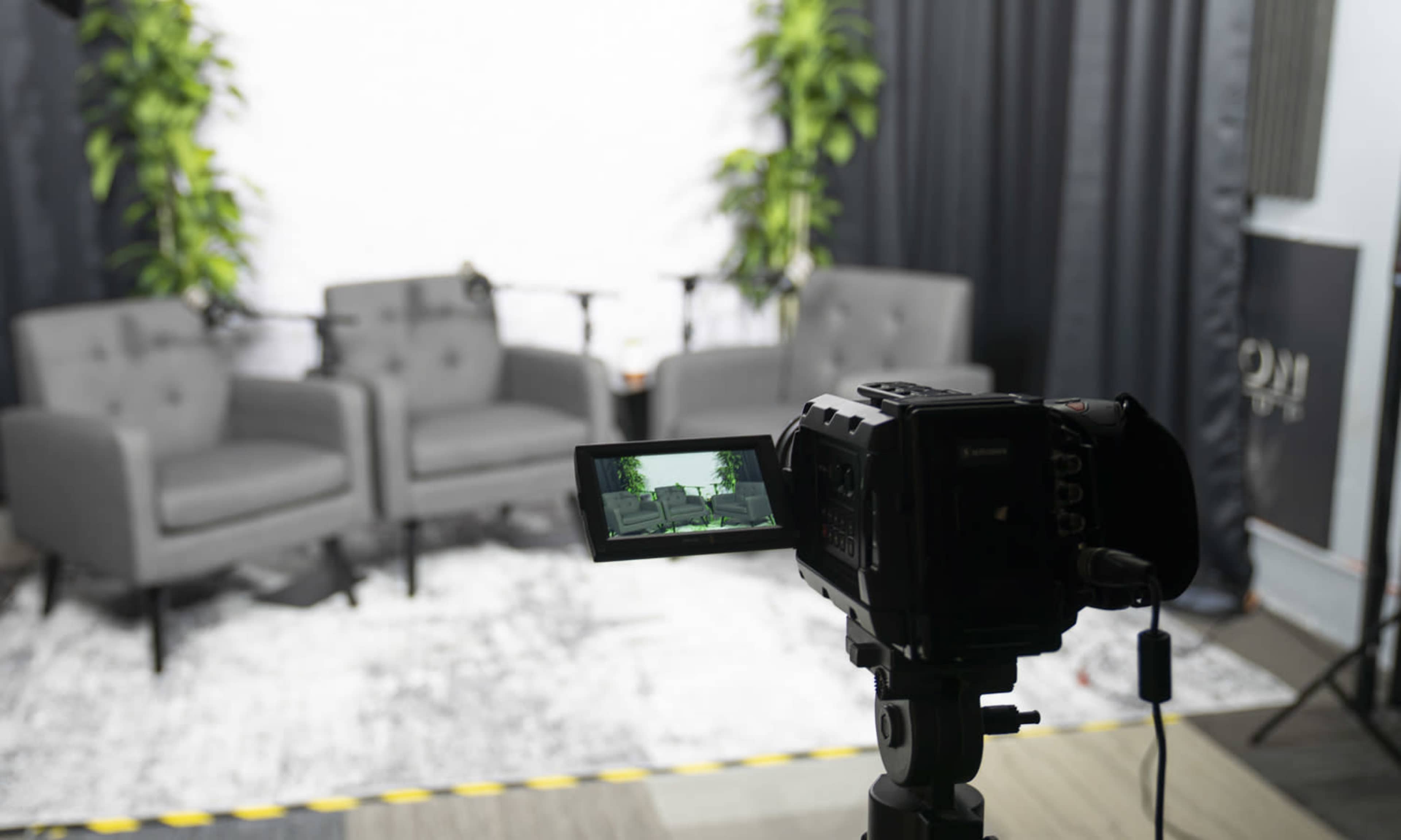 A video camera is positioned in front of two empty gray chairs against a backdrop of indoor plants and a curtain.