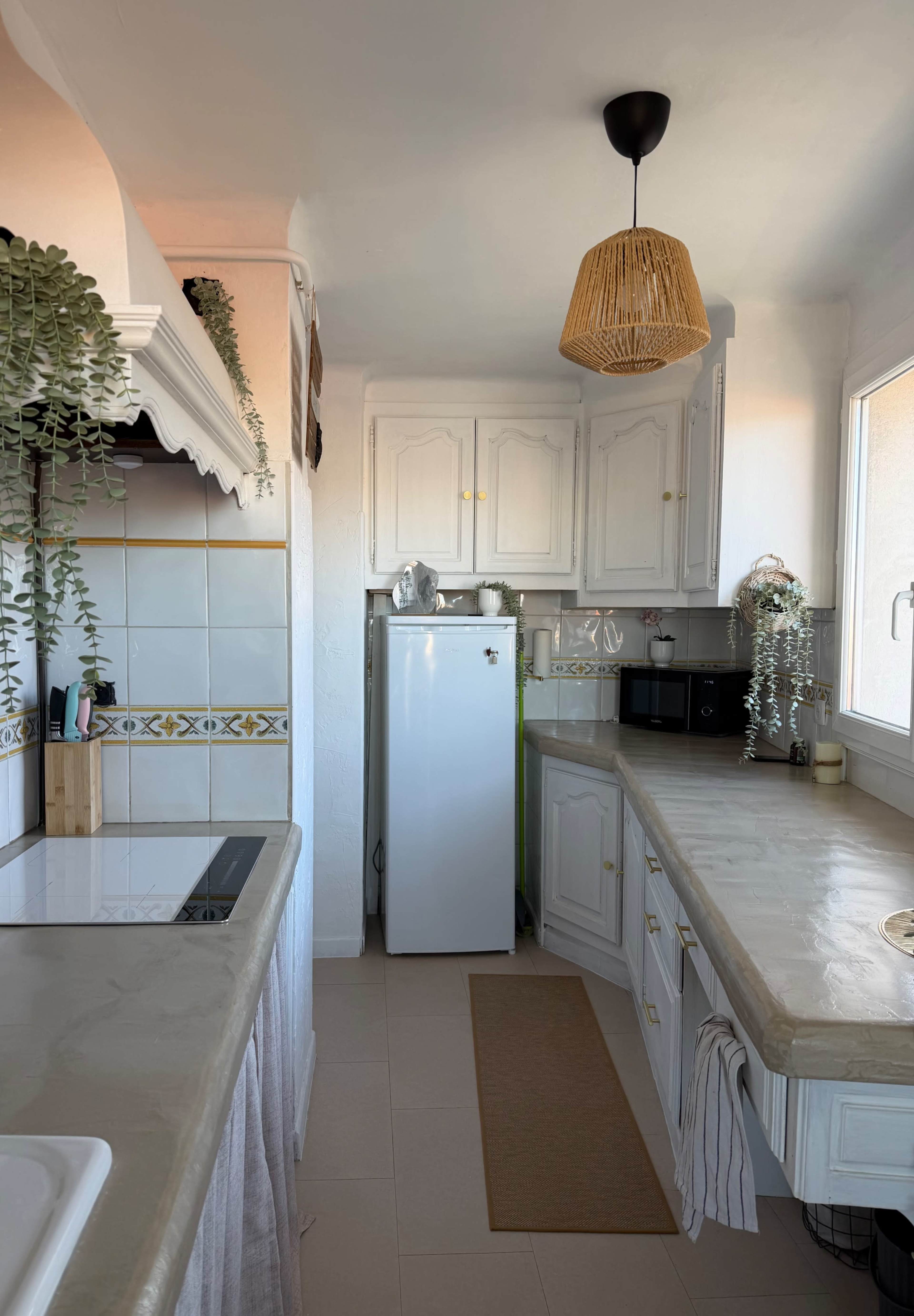 The image shows a bright kitchen with white cabinetry, a refrigerator, and a countertop, featuring natural light and decorative plants.