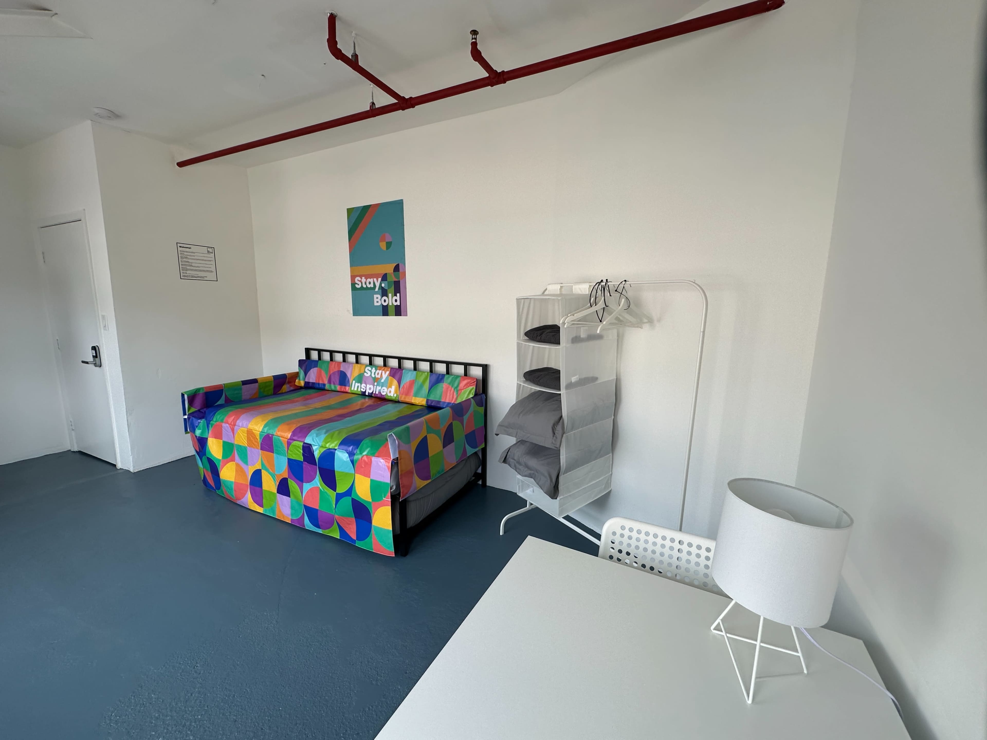 A colorful sofa bed is positioned against a white wall in a bright room, accompanied by a clothing rack, a table, and a lamp.