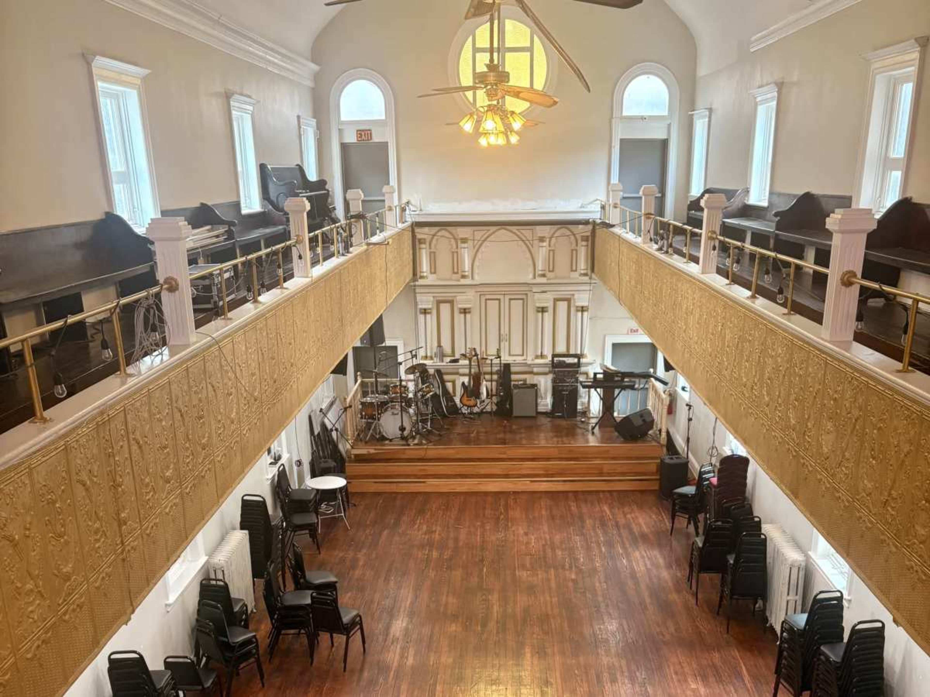 The image shows a spacious, empty hall with wooden floors, a stage at the front, and seating arranged along the sides.