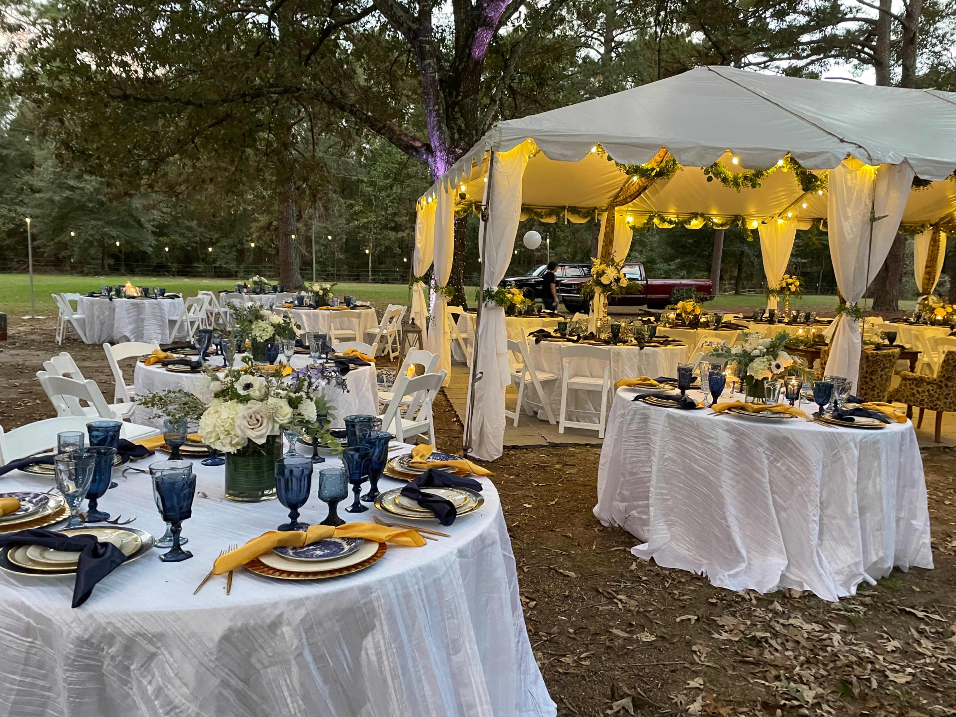 The image shows a decorative outdoor dining setup with white tablecloths, blue glassware, floral centerpieces, and a large covered tent set amidst trees.