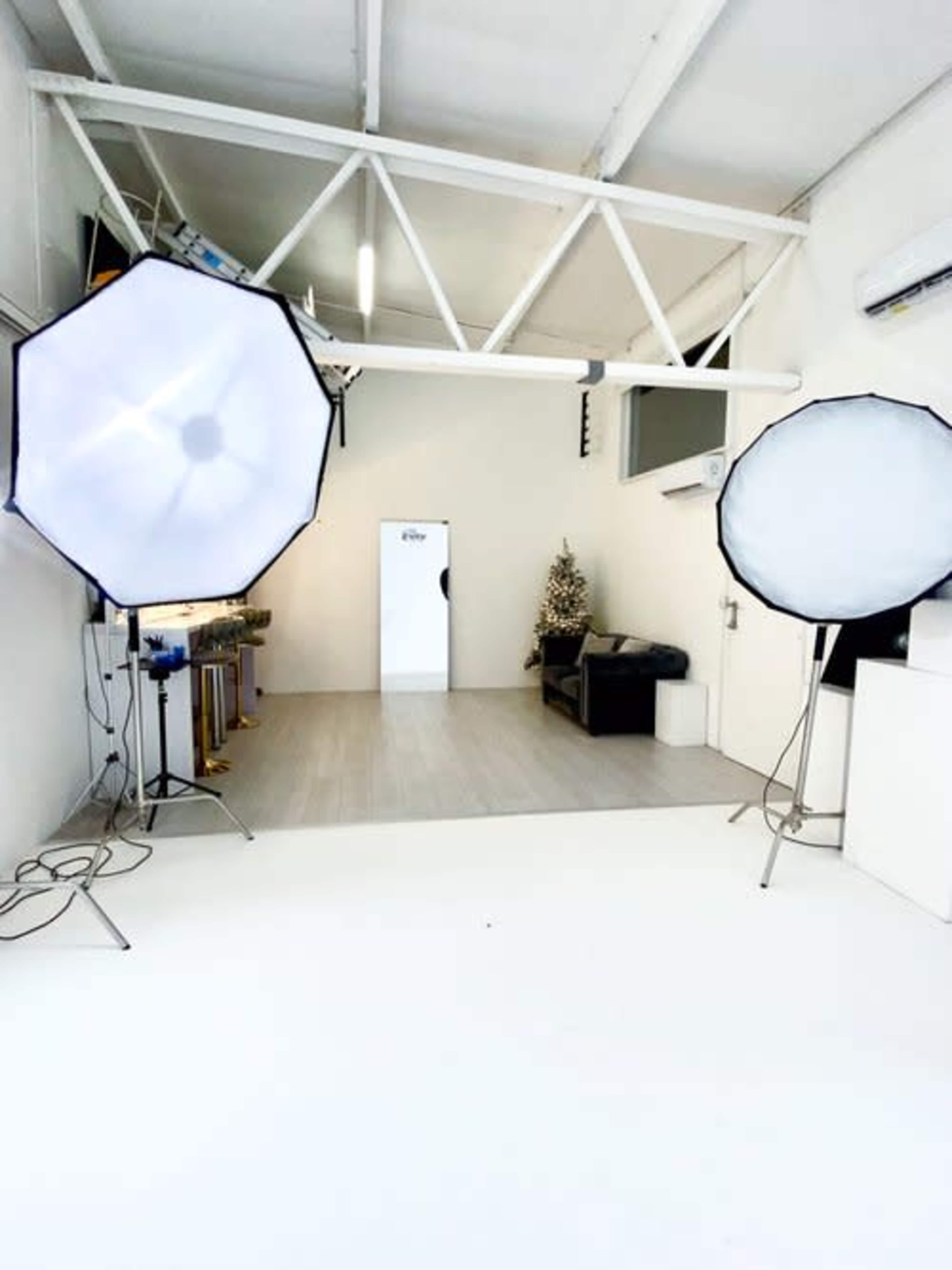 The image shows a well-lit photography studio featuring two softbox lights, a gray sofa, and a white backdrop with a door leading to another room.