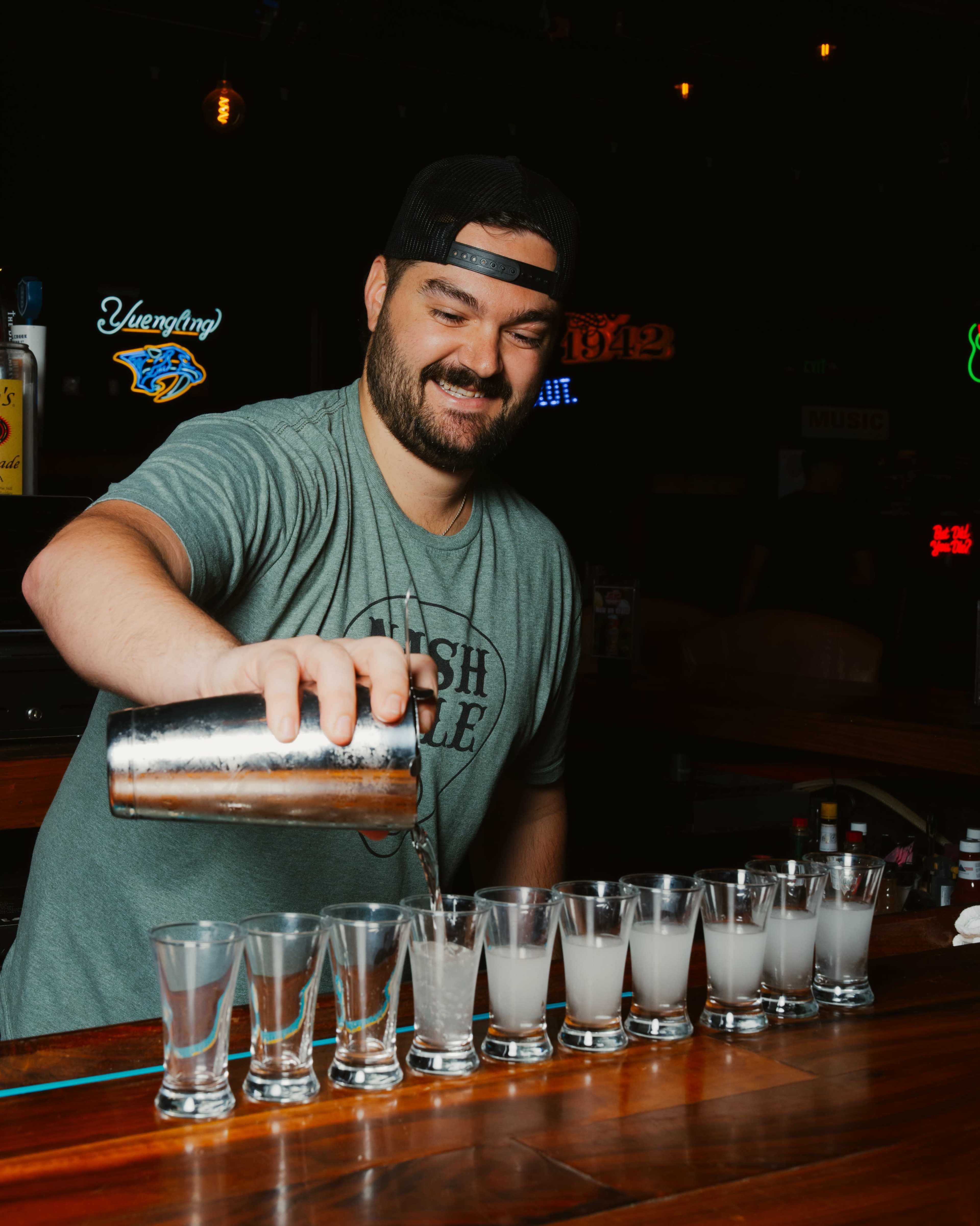 A bartender pours a clear liquid from a shaker into a row of shot glasses on a wooden bar.