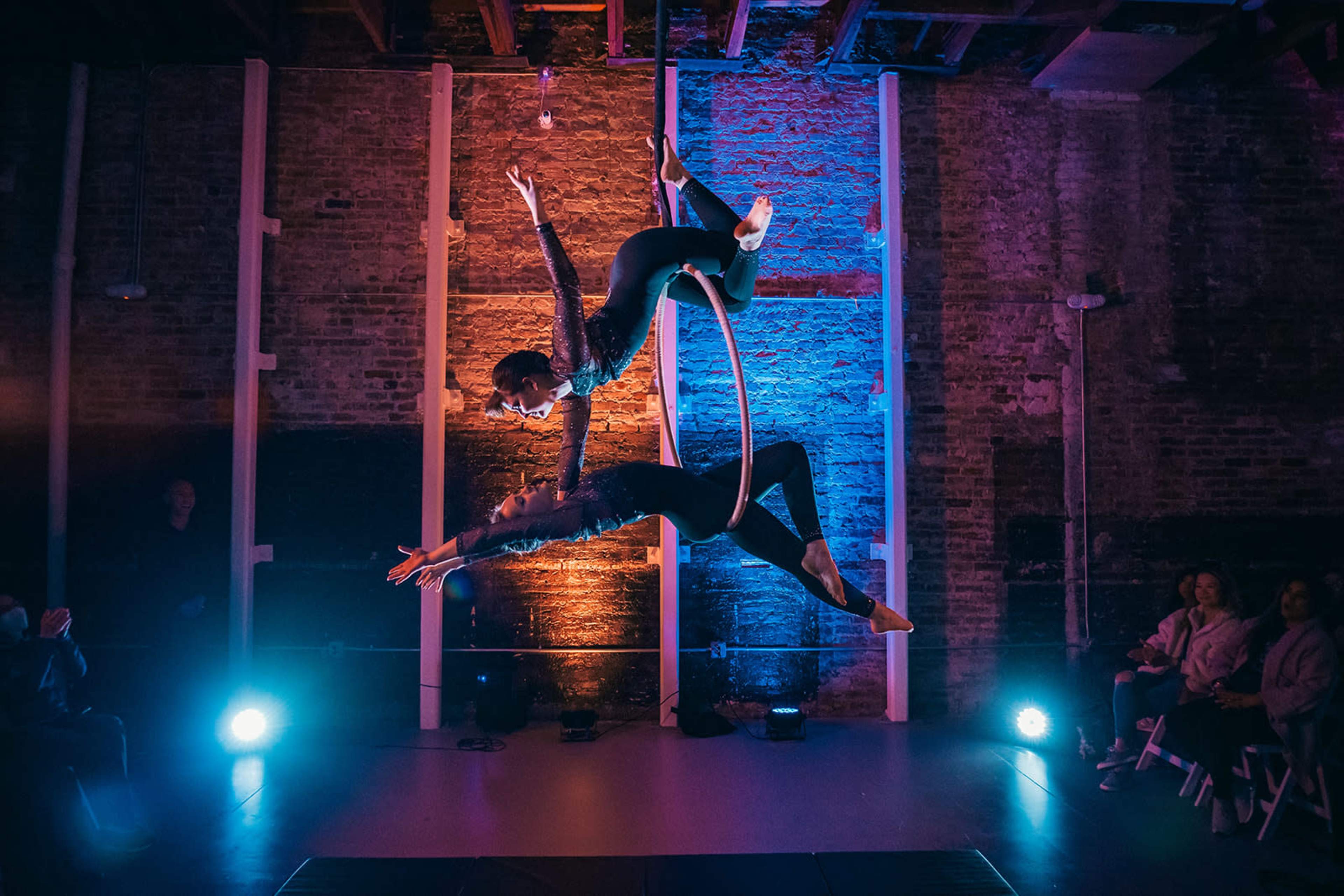 Two acrobats perform an aerial routine on a hoop in a dimly lit space with colorful lighting.