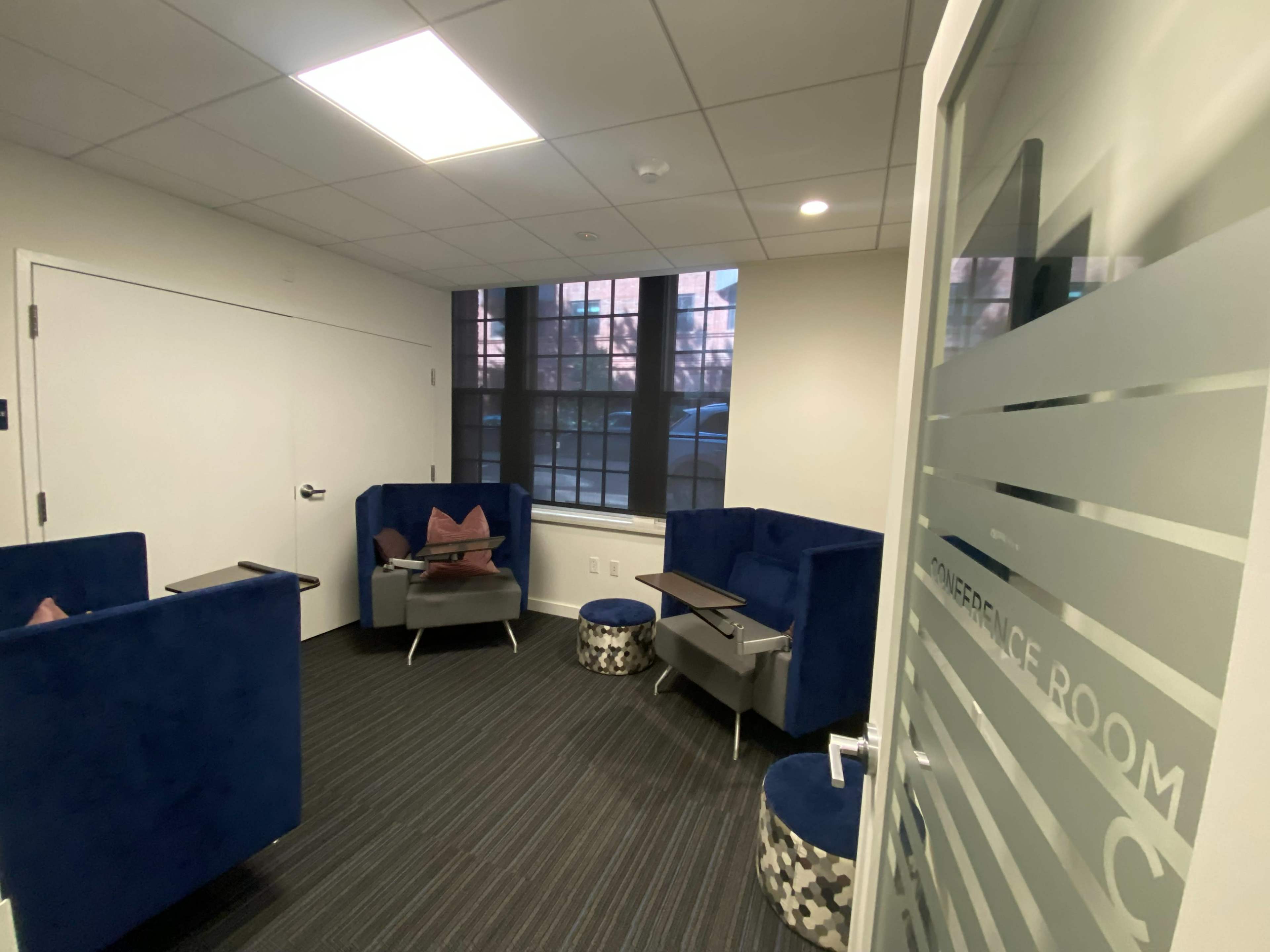 A small conference room entrance featuring blue seating arrangements, a glass door labeled "CONFERENCE ROOM," and a view of a window with blinds.