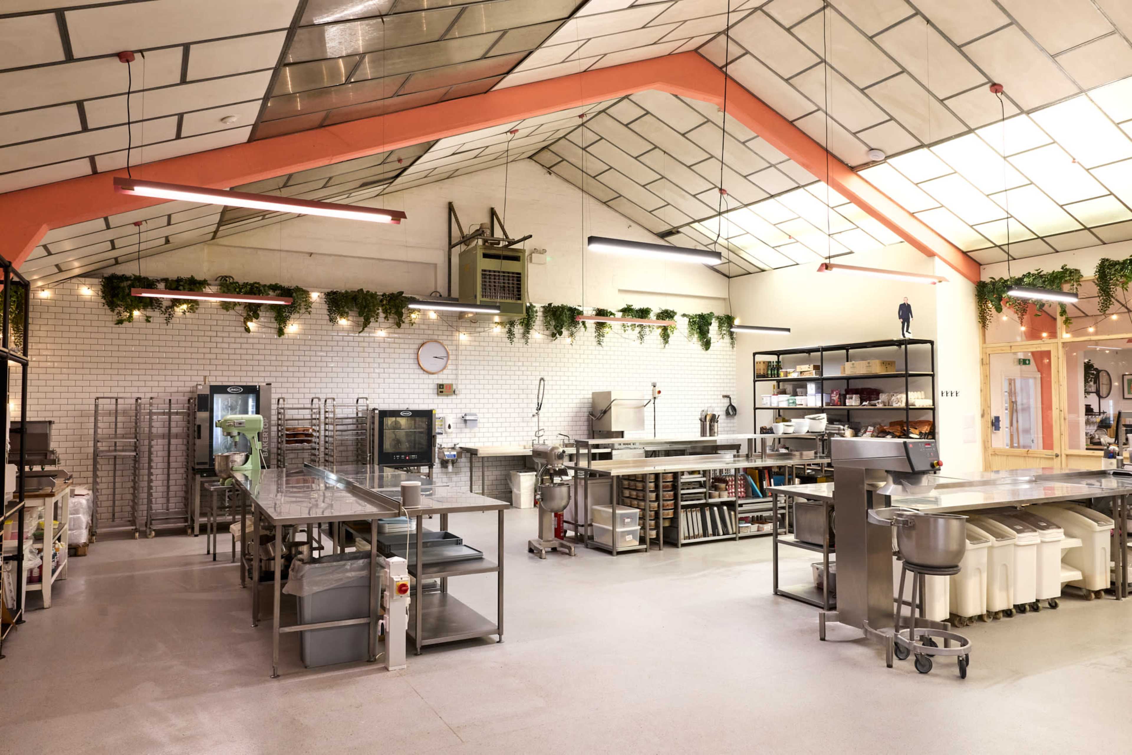 The image shows a spacious industrial kitchen with metal worktables, various kitchen appliances, and overhead lighting, all set under a skylight.
