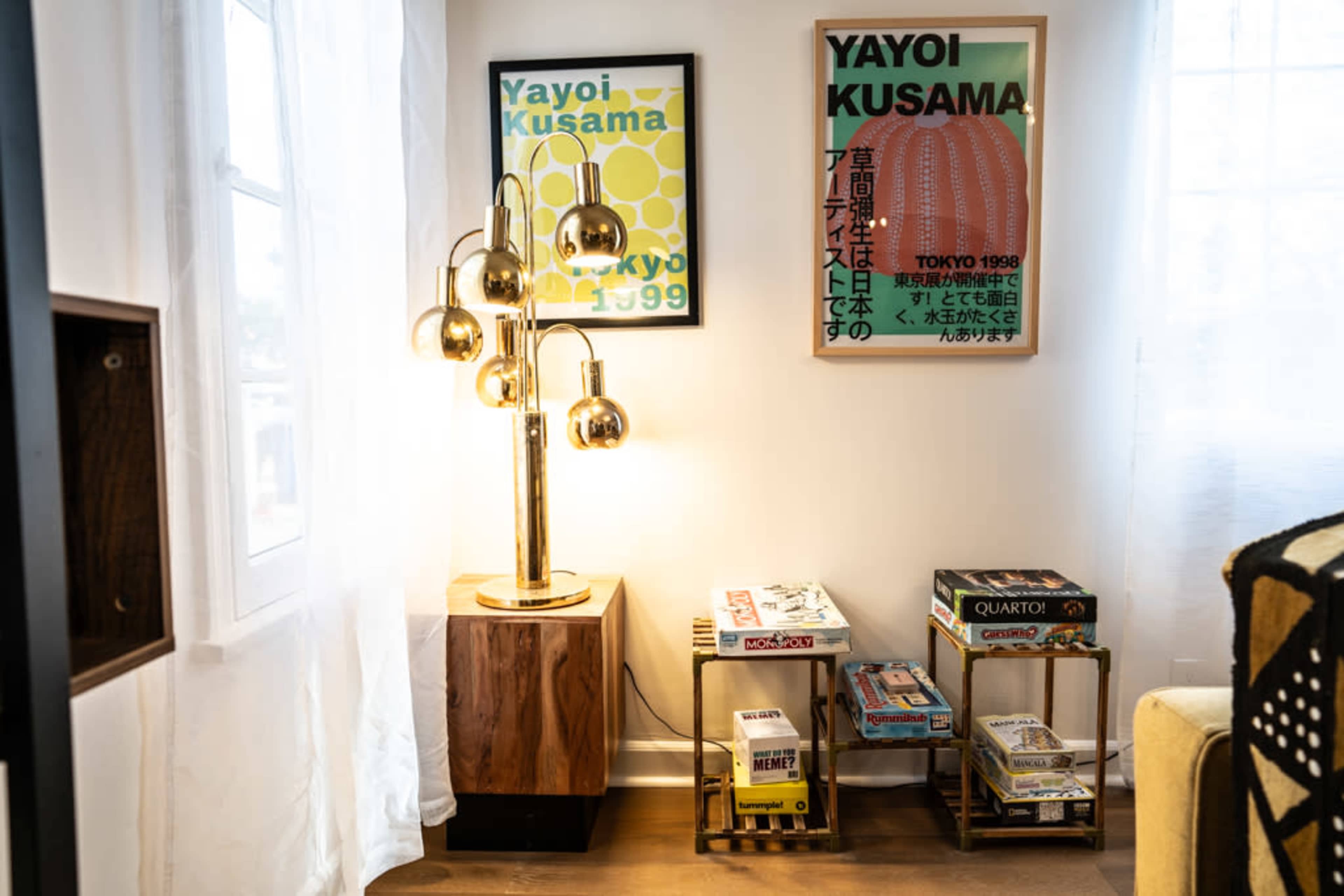 The image shows a cozy living room corner featuring a gold floor lamp, two framed posters of Yayoi Kusama, and a small bookshelf with various board games.