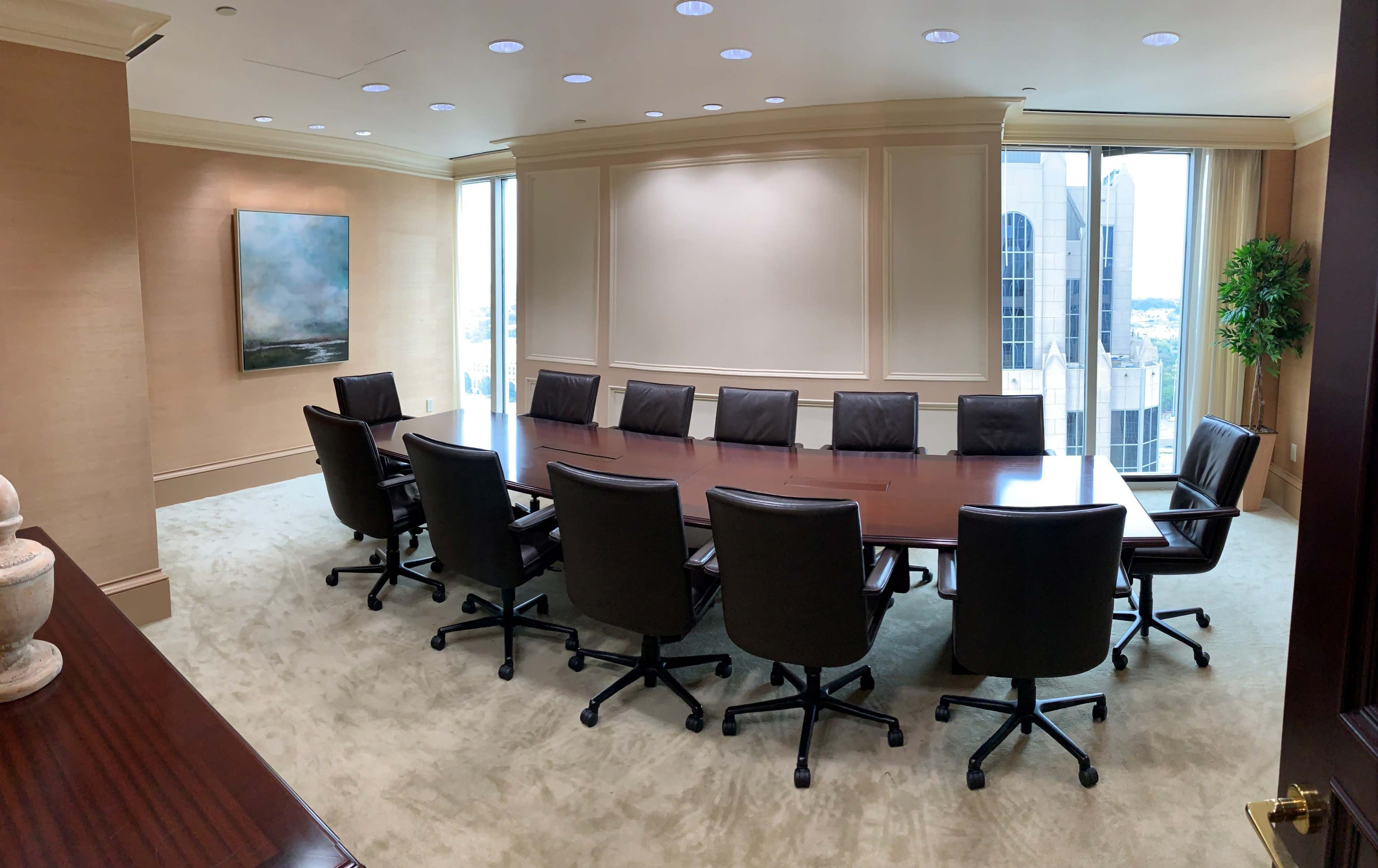 A conference room features a long wooden table surrounded by black leather chairs, with large windows displaying a city skyline.