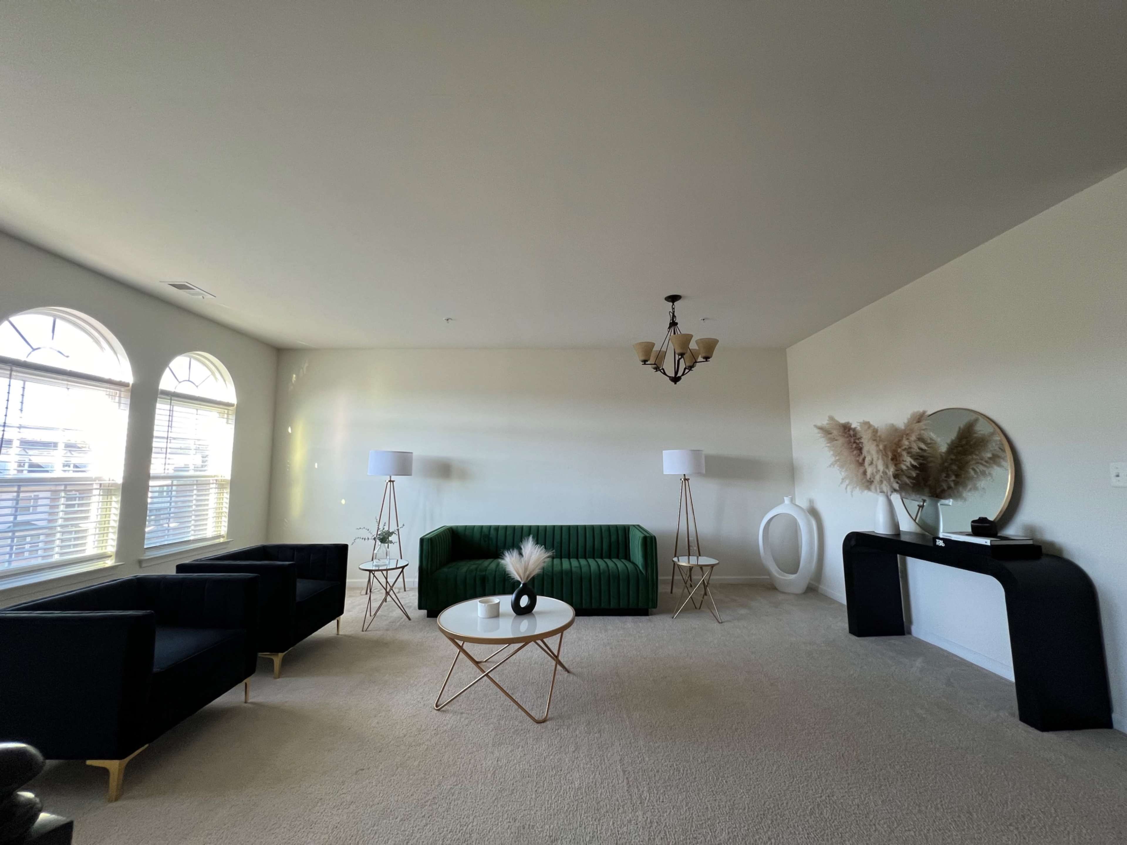 A spacious living room features a green sofa, two black armchairs, a round coffee table, and a console table adorned with decorative elements, under a chandelier and natural light from large windows.