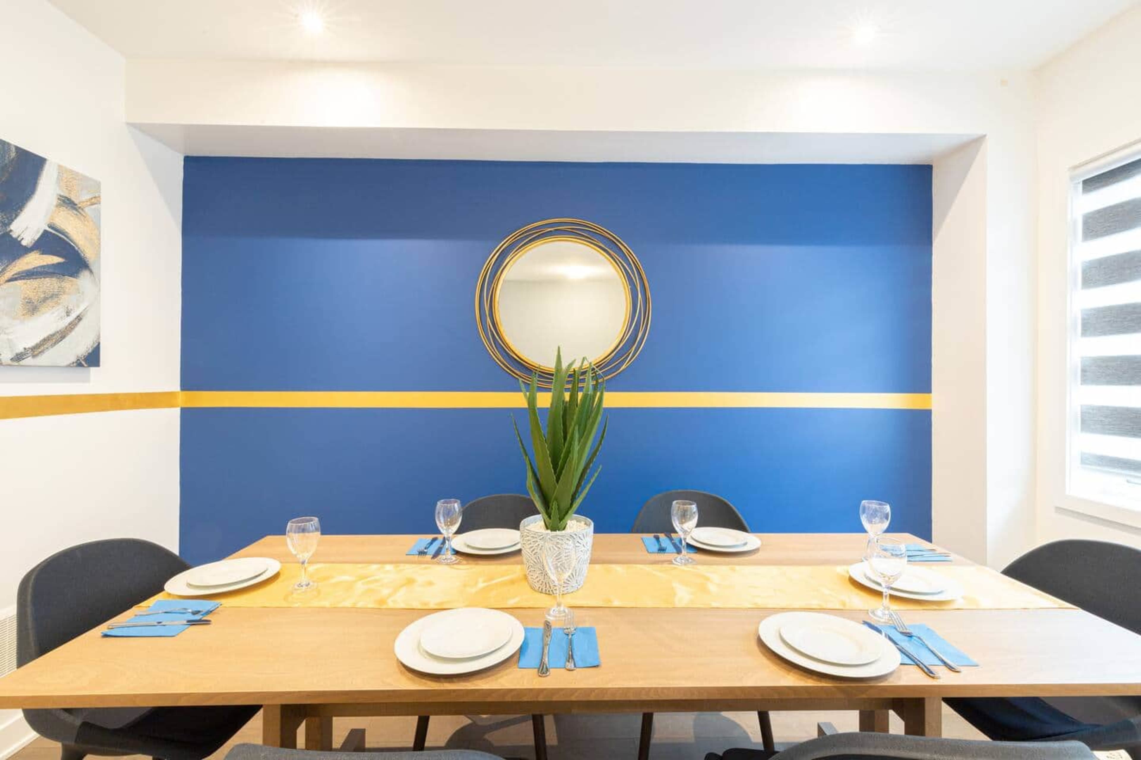 A dining table is set with plates and glasses in a room featuring a blue accent wall with a circular mirror and a potted plant centerpiece.