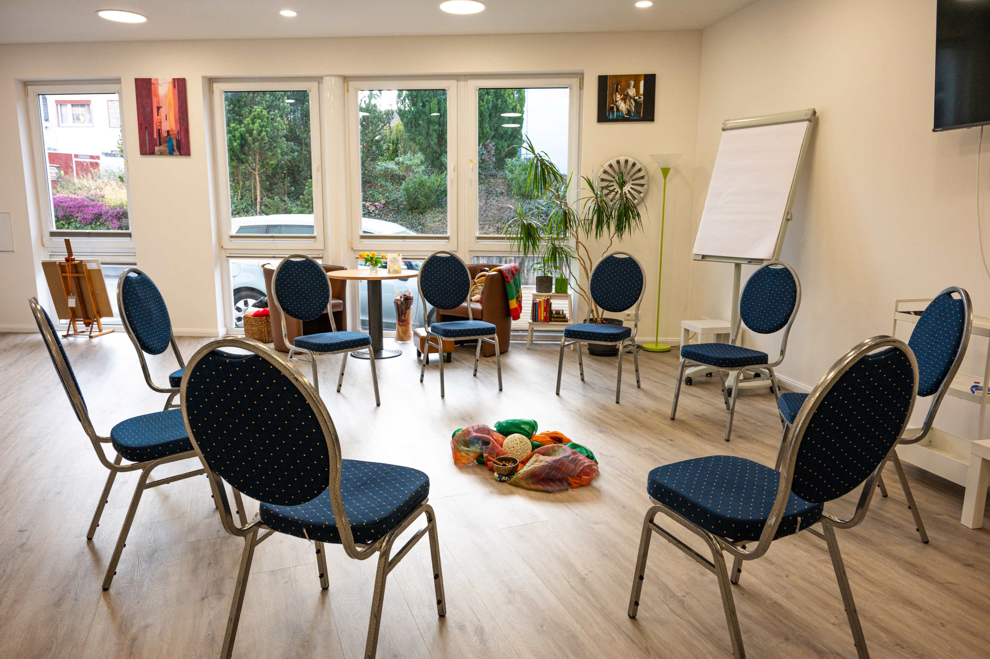 A circle of blue chairs is arranged around a colorful assortment of objects on the floor in a well-lit room with large windows.