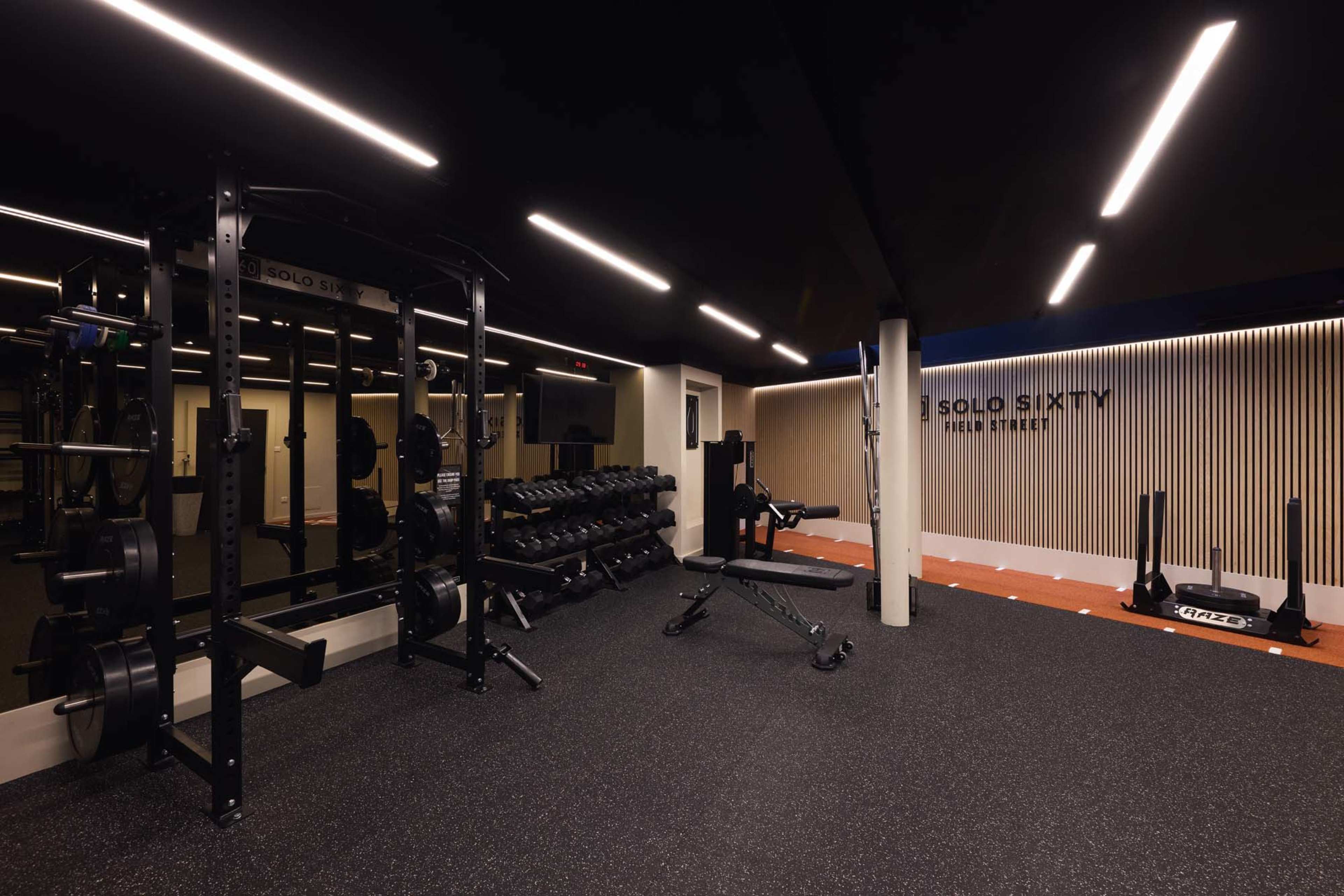 The image depicts a modern gym space with weightlifting equipment, including a power rack, dumbbell rack, and a bench, under bright ceiling lights.