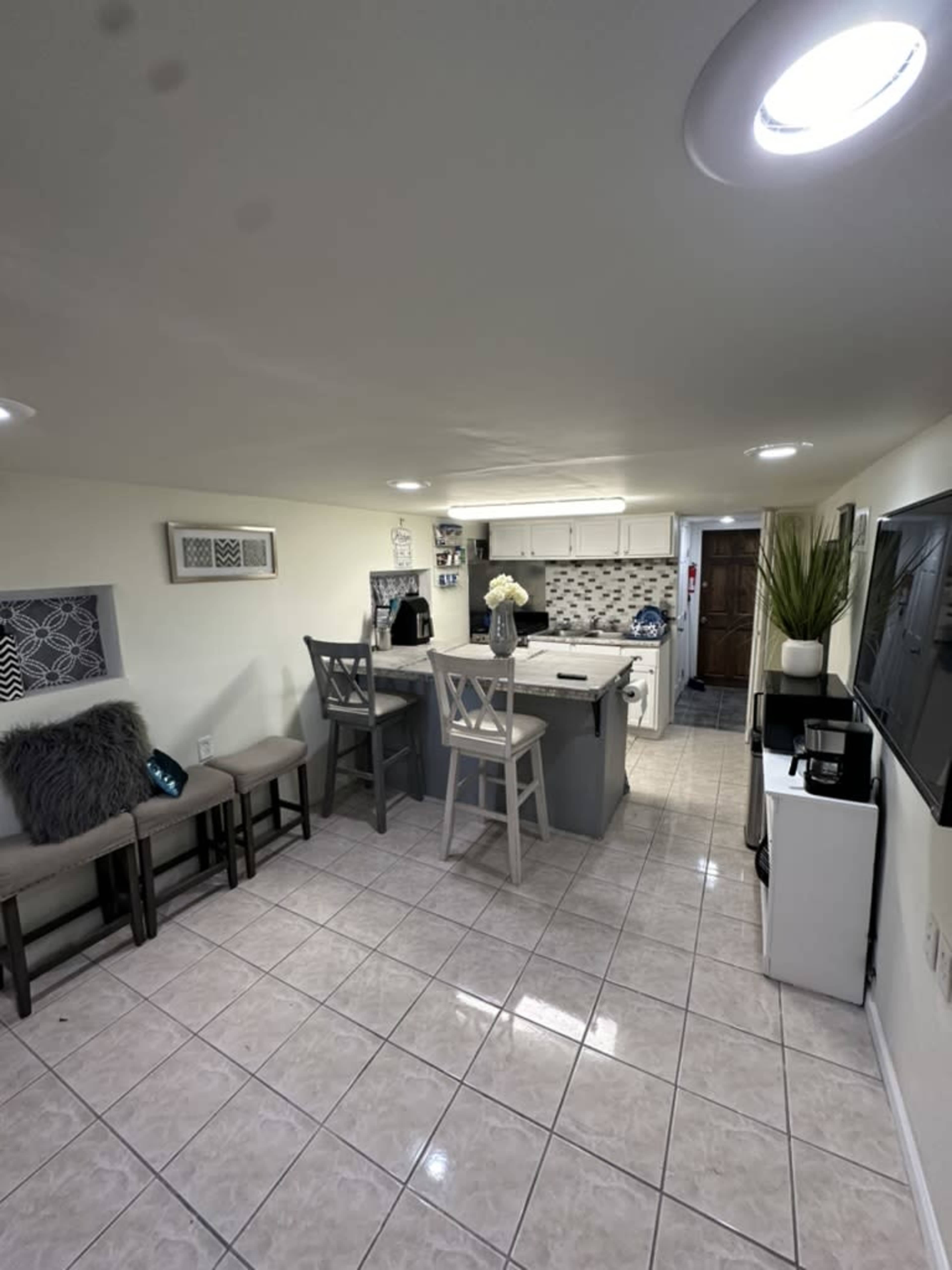 The image shows a small, modern kitchen area with a dining table and chairs, light tile flooring, and a variety of kitchen appliances.