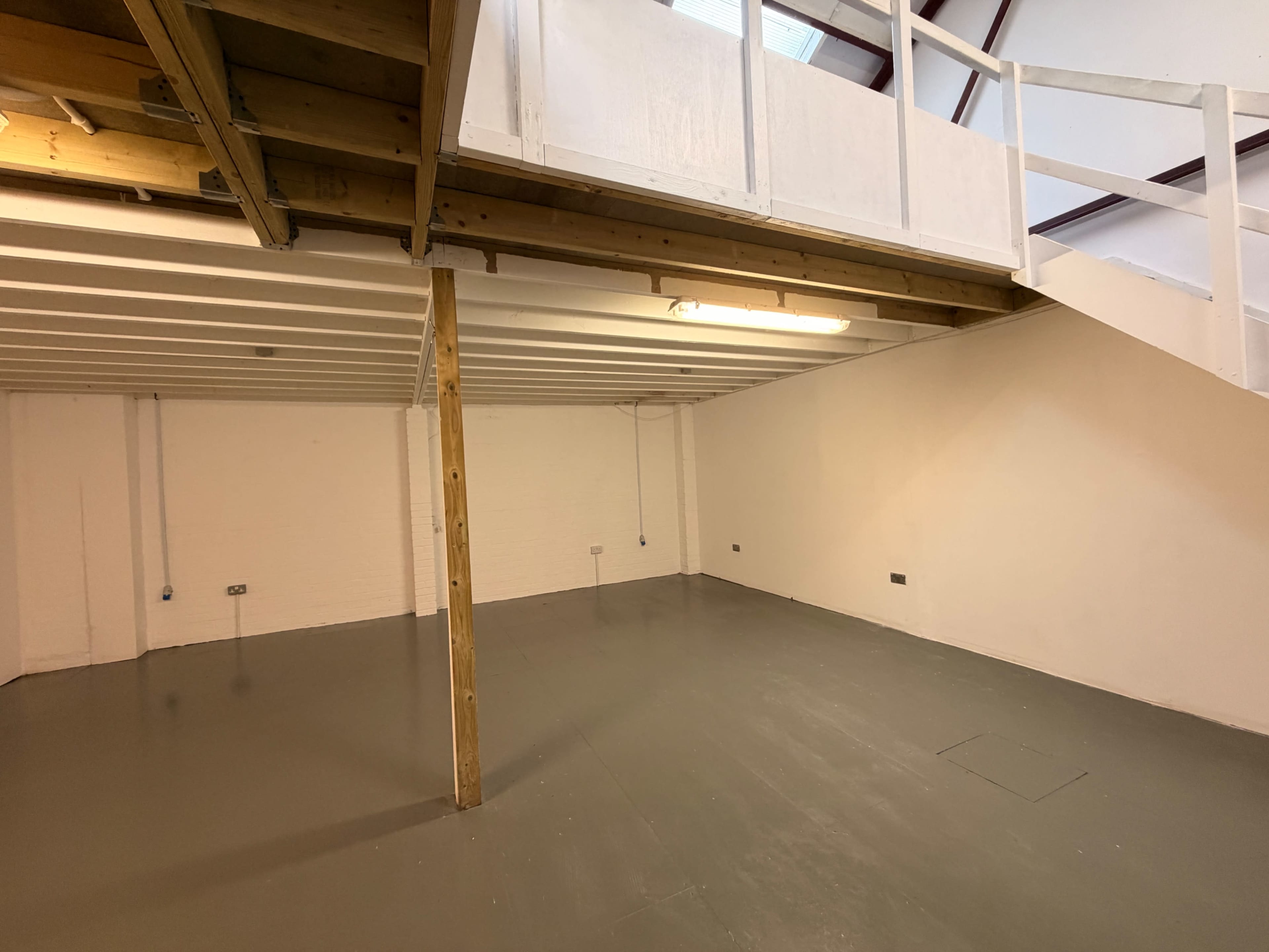 The image shows an interior room with a wooden beam supporting a mezzanine level, featuring a bare concrete floor and white walls.