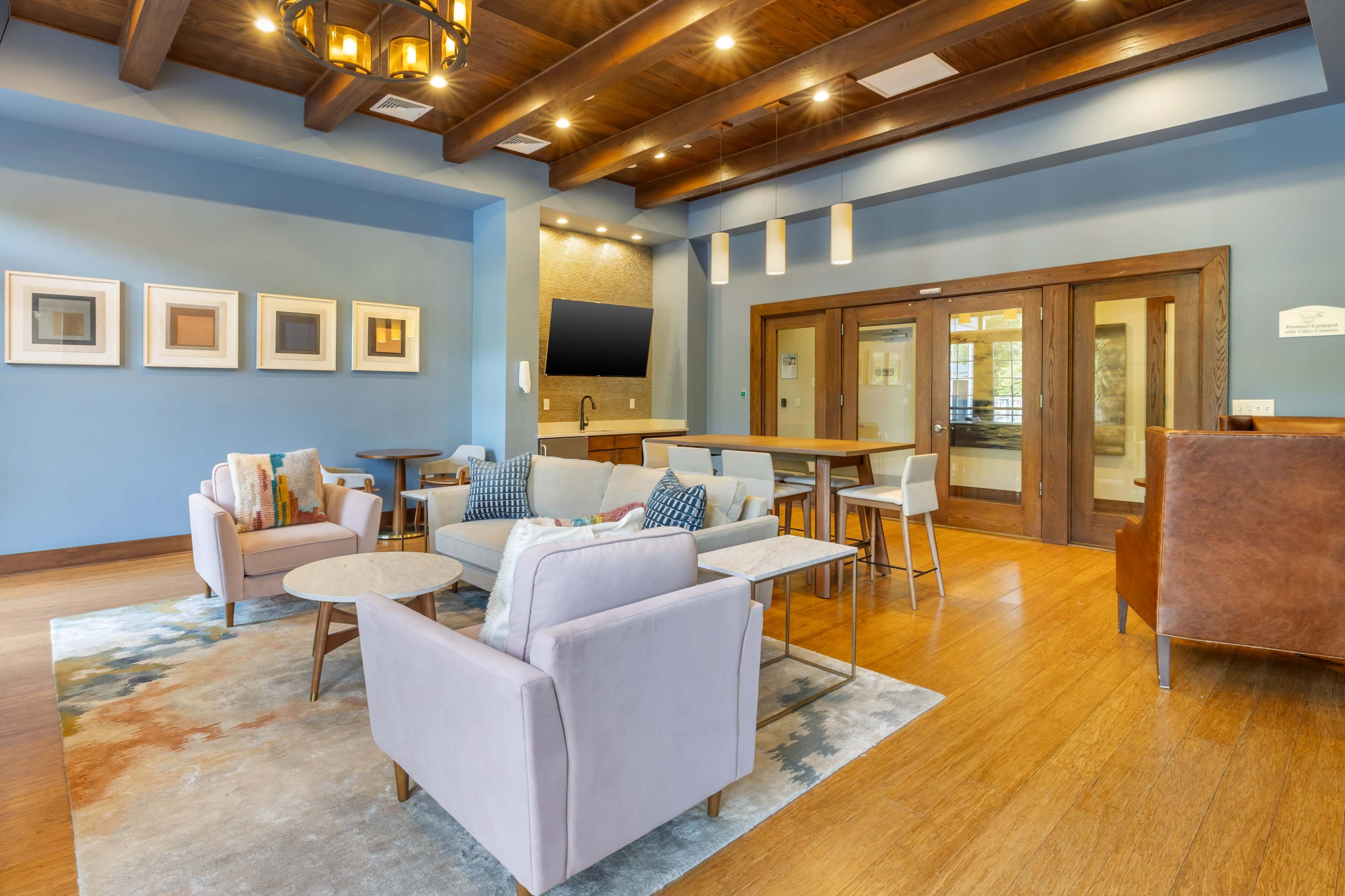 The image shows a modern lobby area featuring comfortable seating, tables, and a wall-mounted television, set against a backdrop of blue and wooden tones.