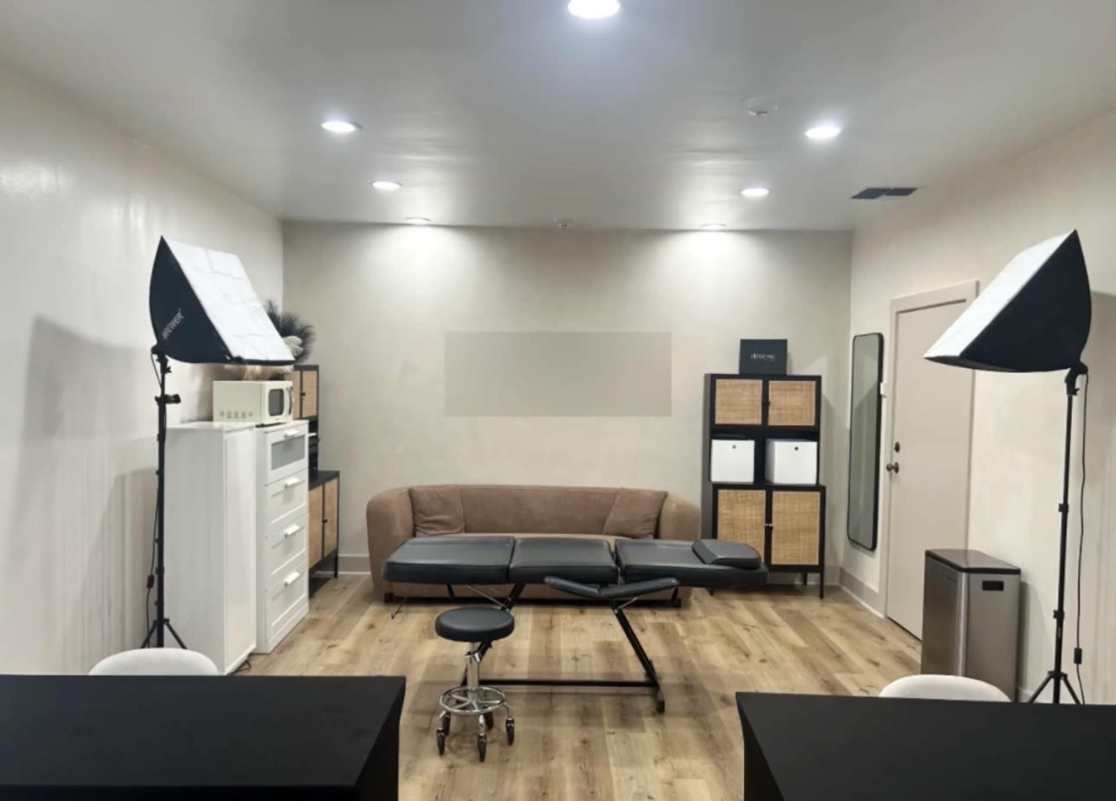 The image shows a clean, well-lit room featuring a brown sofa, a treatment table, two softbox lights, and storage units along the walls.