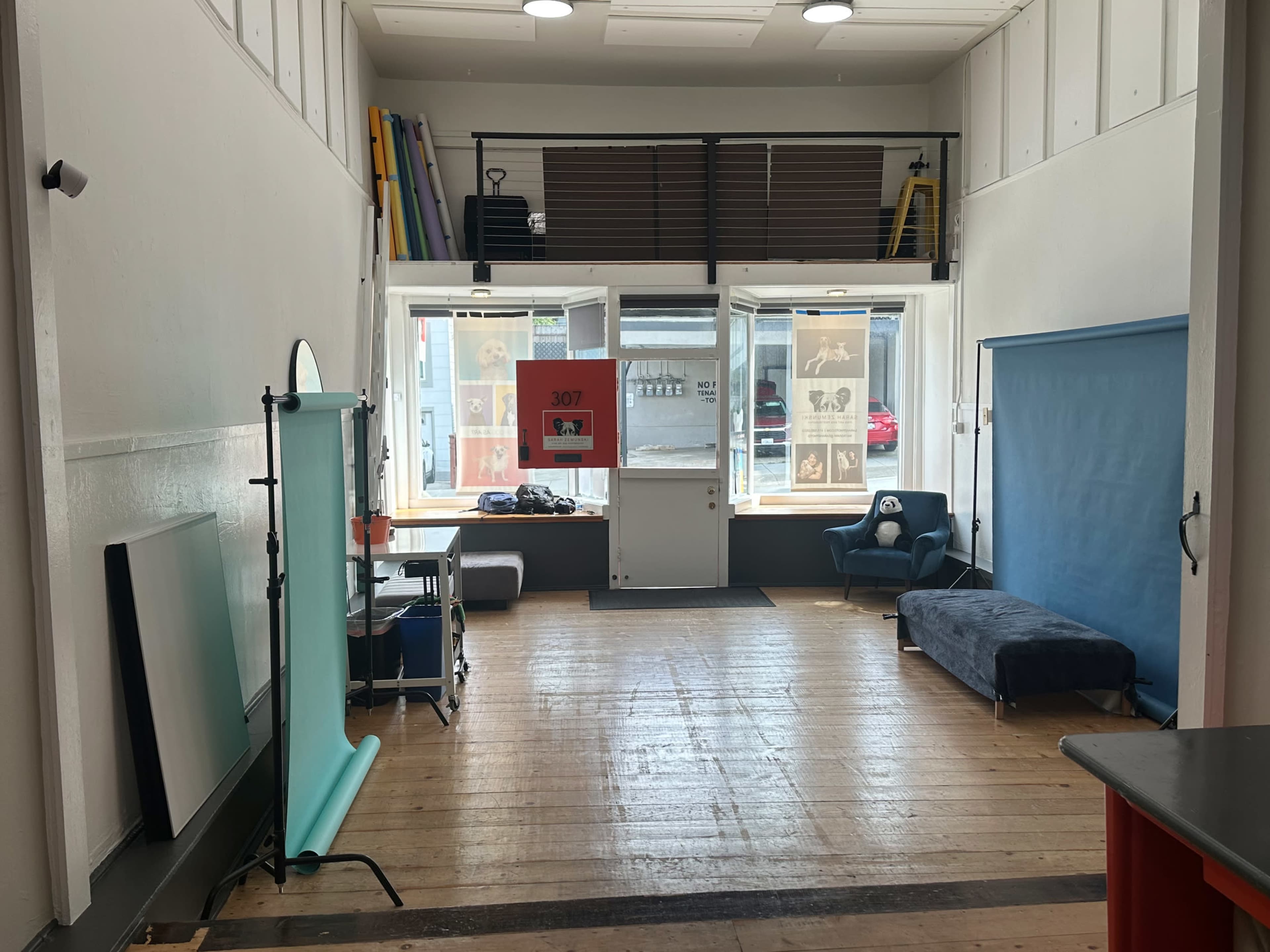 The image shows the interior of a spacious studio with wooden floors, large windows, and various photography equipment, including a backdrop and seating area.