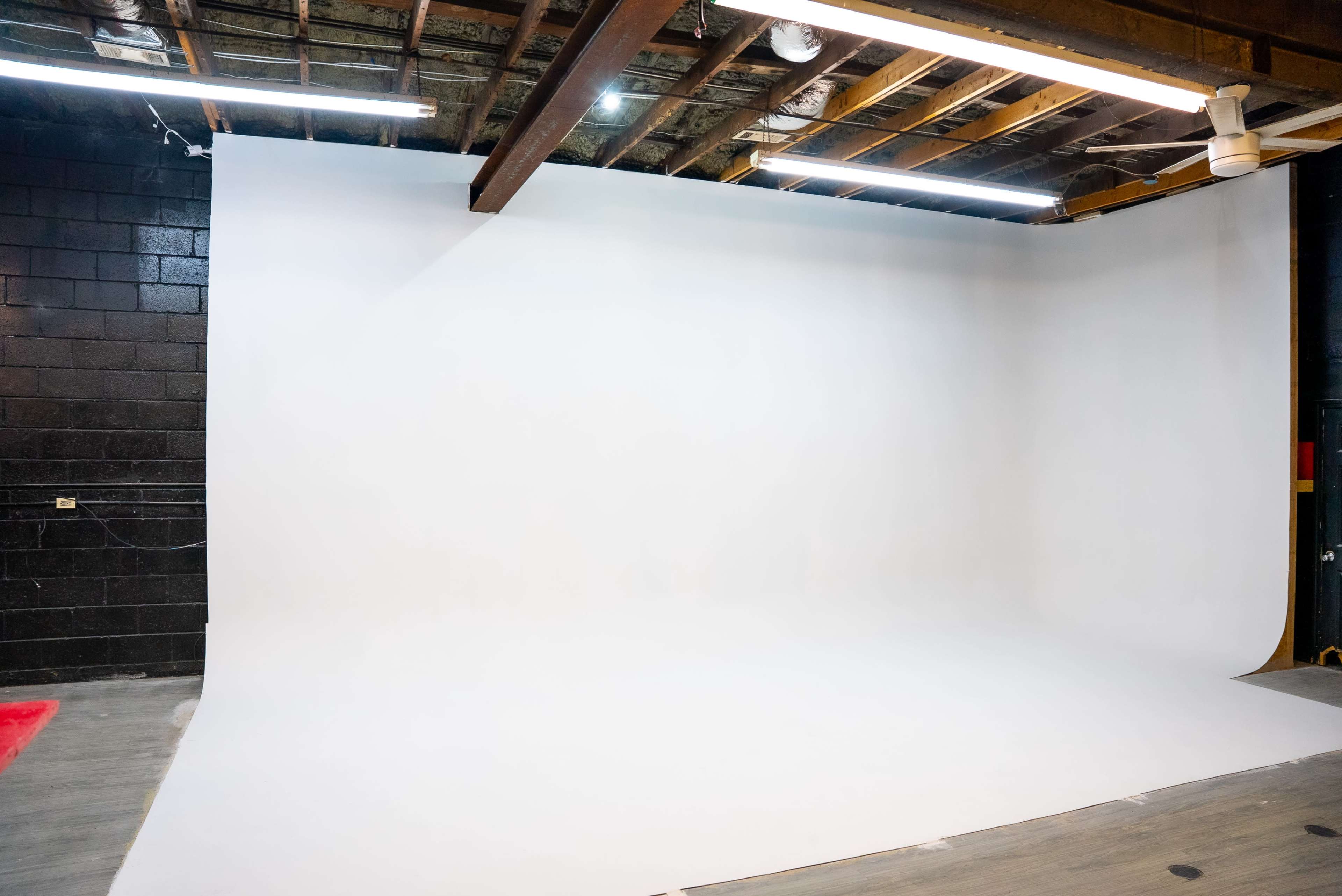 The image shows a blank photography studio with a white seamless backdrop and exposed wooden beams above.