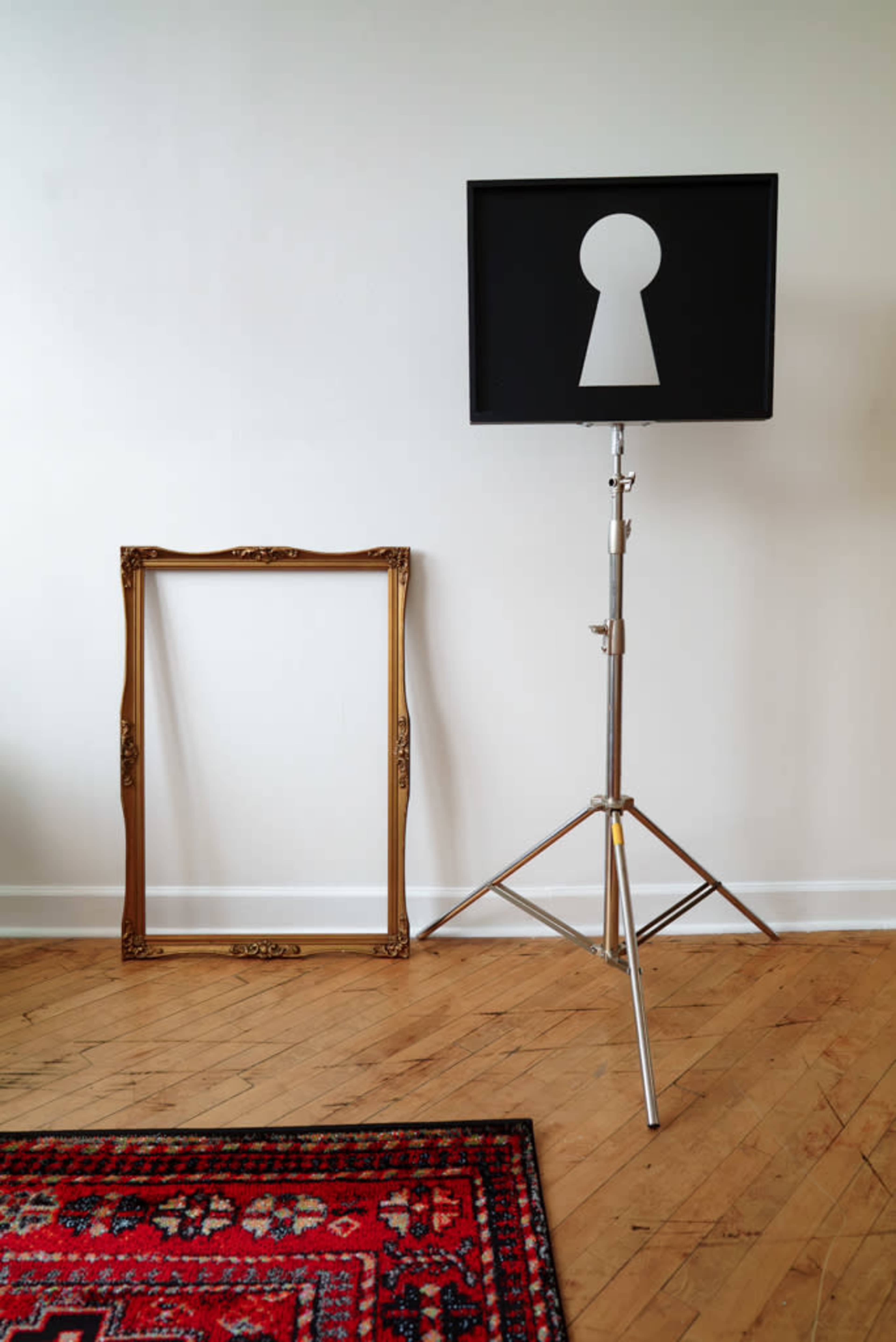 The image features a black box resembling a keyhole on a tripod stand, positioned next to an empty gold-framed picture frame, all set against a plain white wall and wooden floor.
