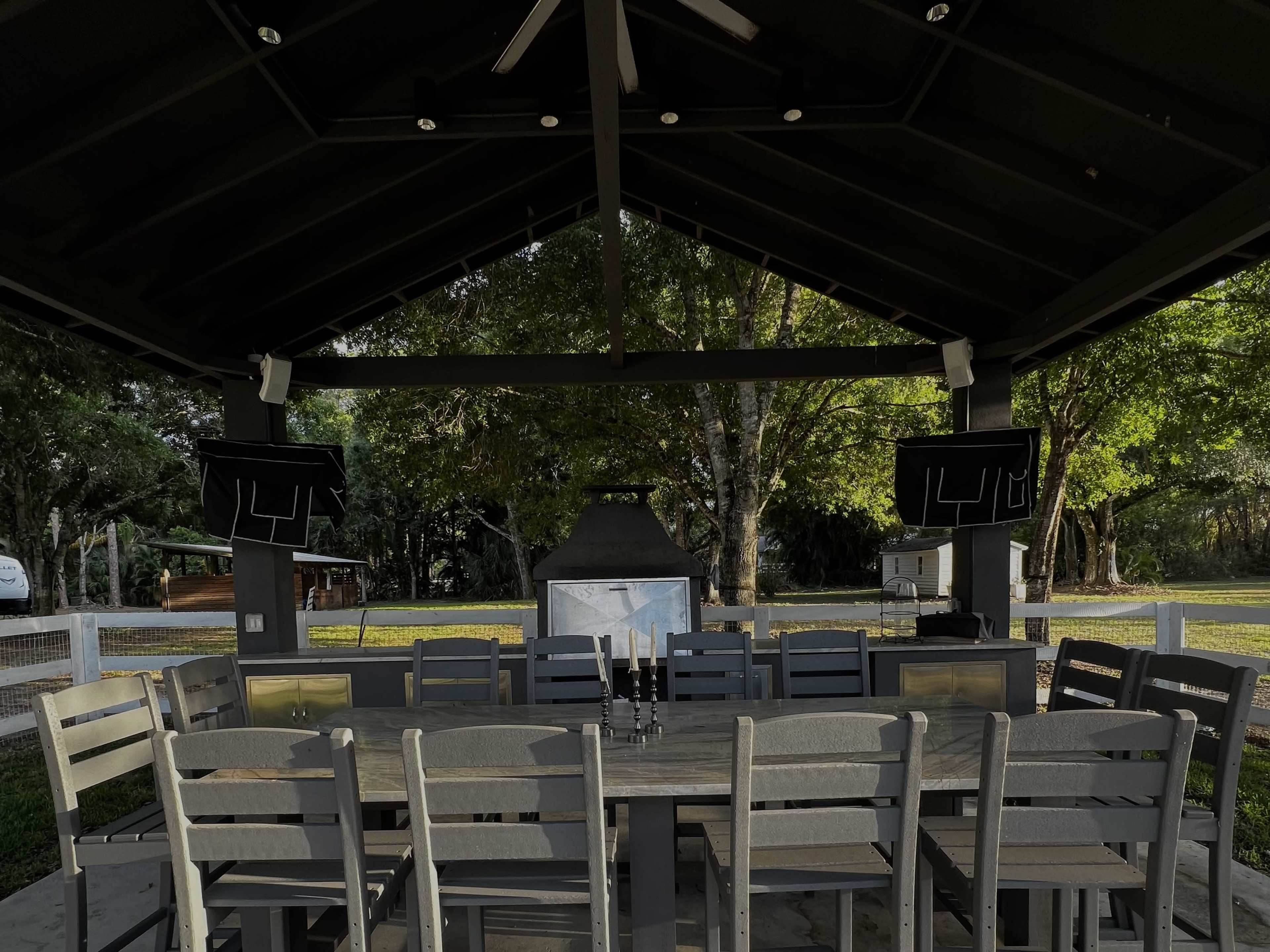 A spacious outdoor pavilion features a long wooden dining table surrounded by chairs, with an outdoor grill and two mounted screens in the background.