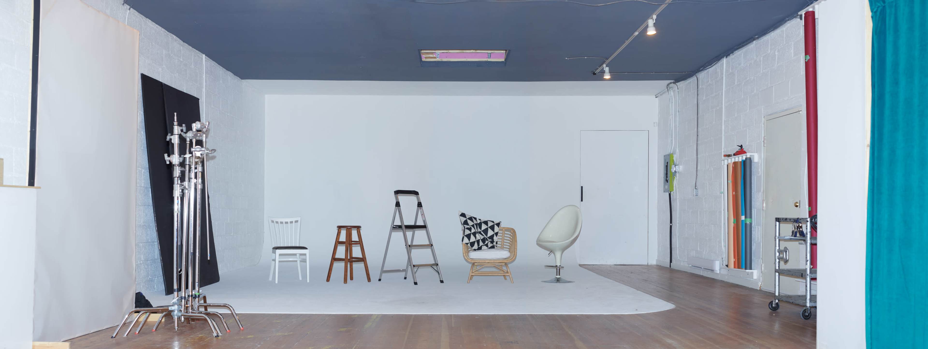 A spacious photography studio with a wooden floor, white backdrop, and several chairs and stools arranged in the center.