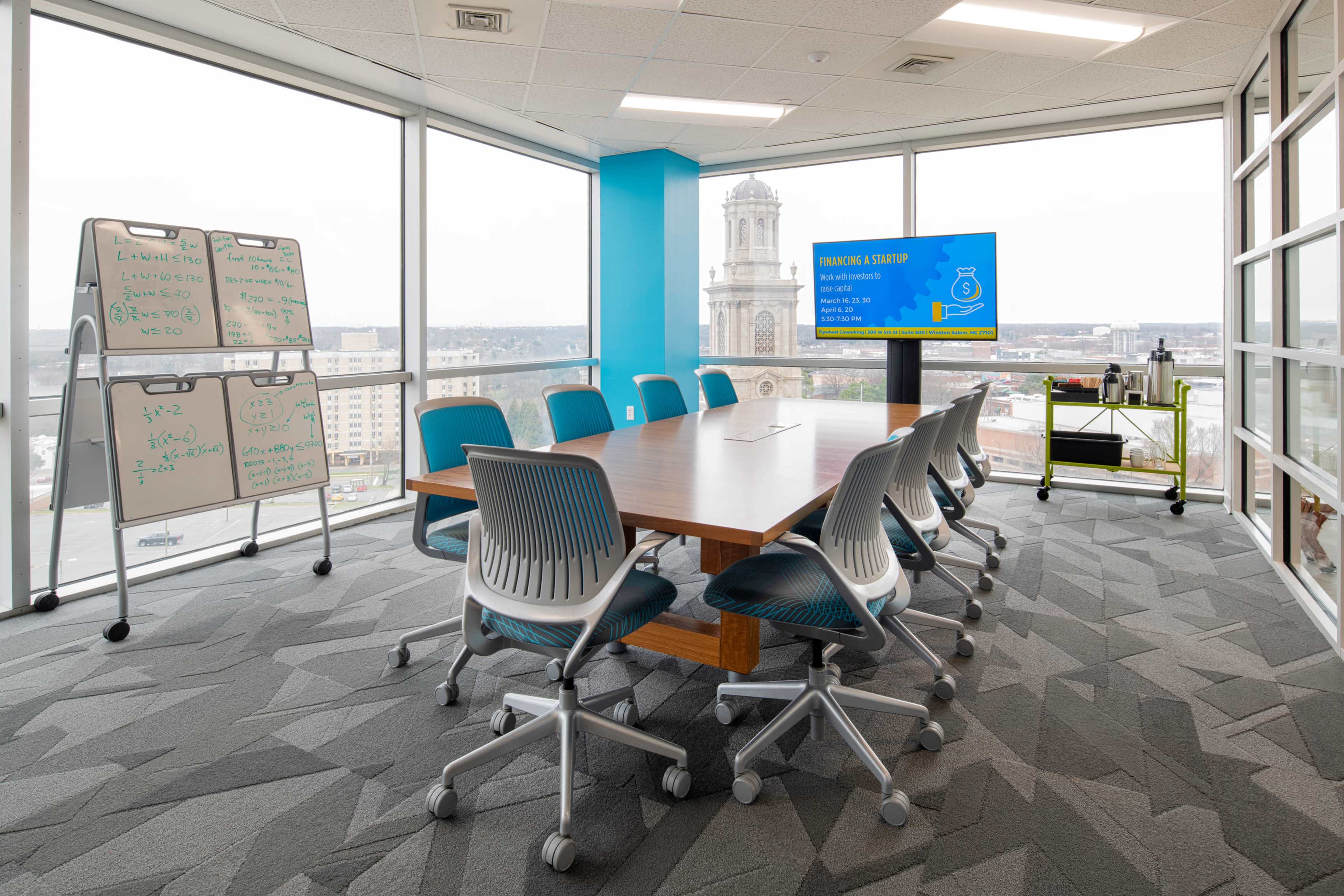 A modern conference room features a large wooden table surrounded by ergonomic chairs, with a screen displaying information and whiteboards for brainstorming, all in a sunlit space with views of the city outside.