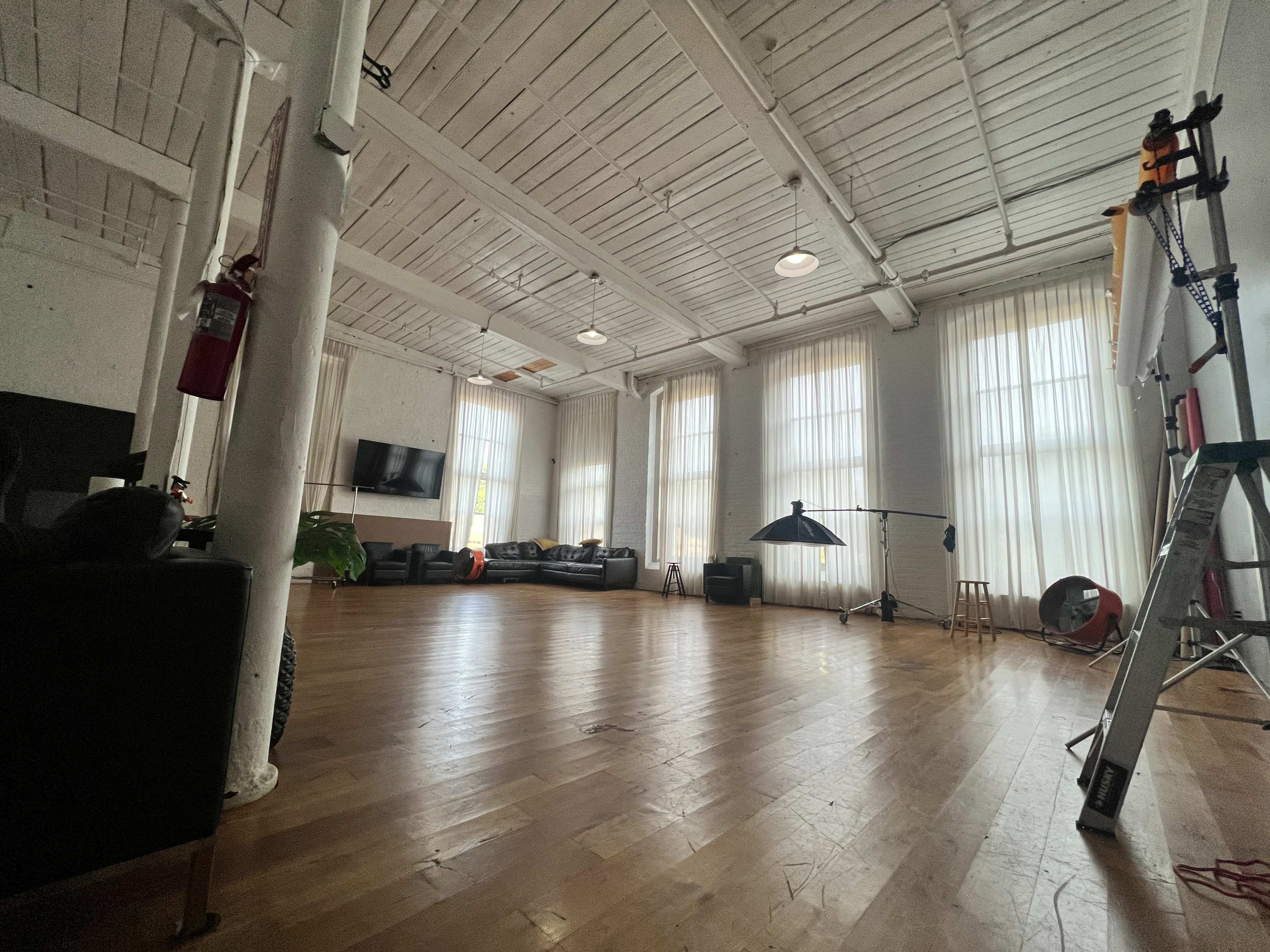 The image shows a large, open studio space with wooden floors, tall windows covered by sheer curtains, a black couch, and photography equipment in the corner.