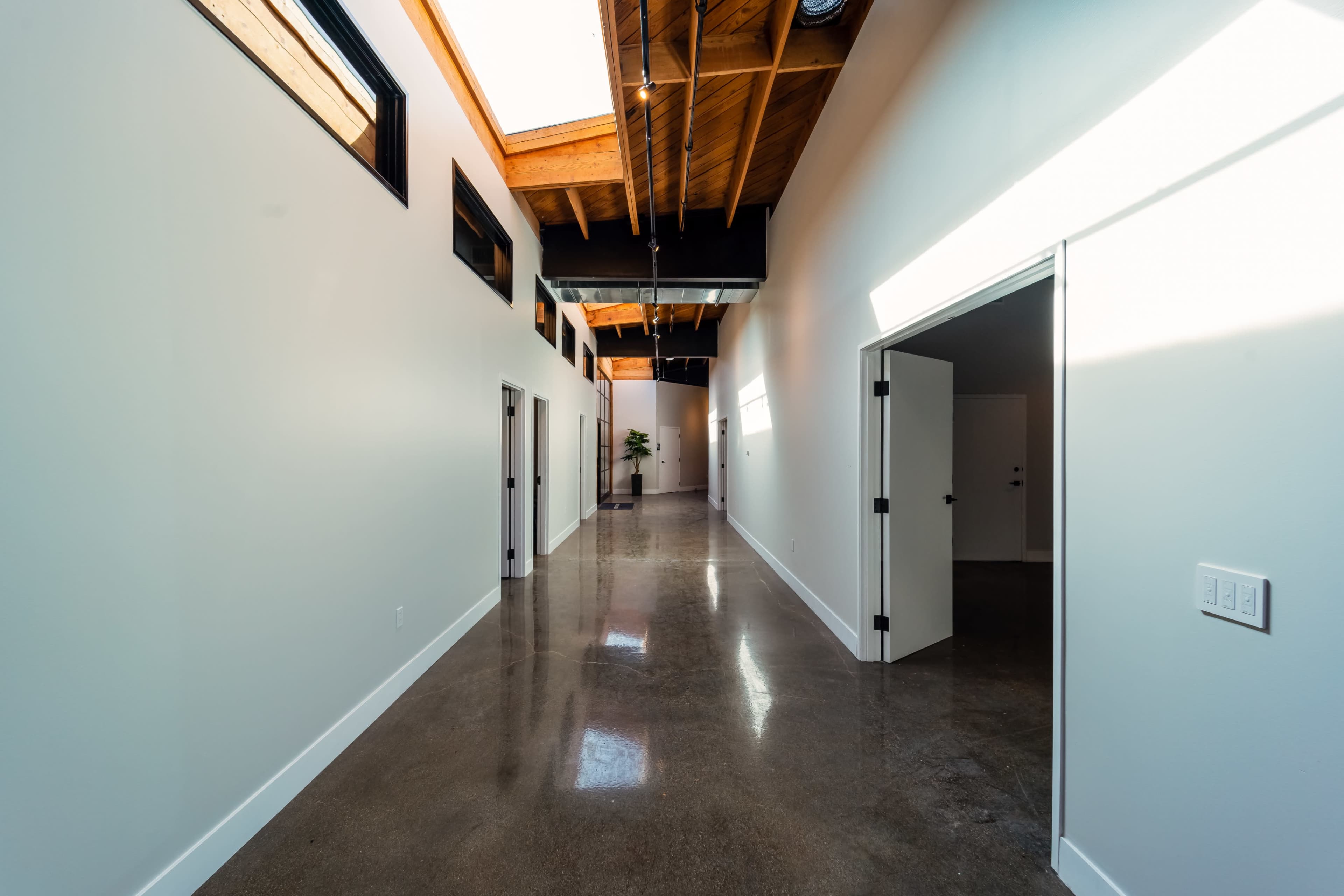The image shows a modern corridor with polished concrete flooring, high ceilings with exposed wooden beams, and multiple doors along the walls.