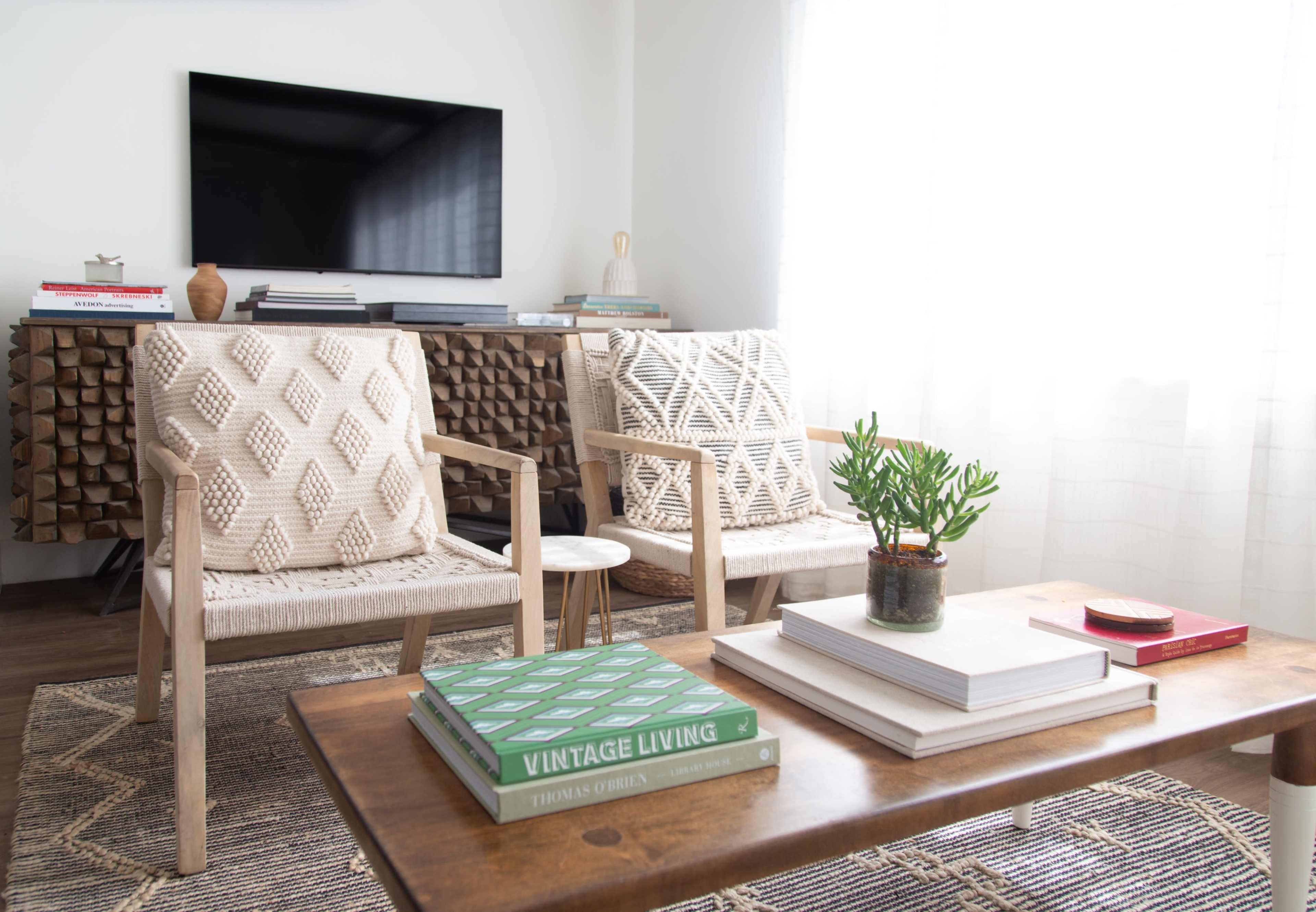 The image shows a modern living room with two patterned chairs, a wooden coffee table stacked with books, and a large television mounted on the wall.
