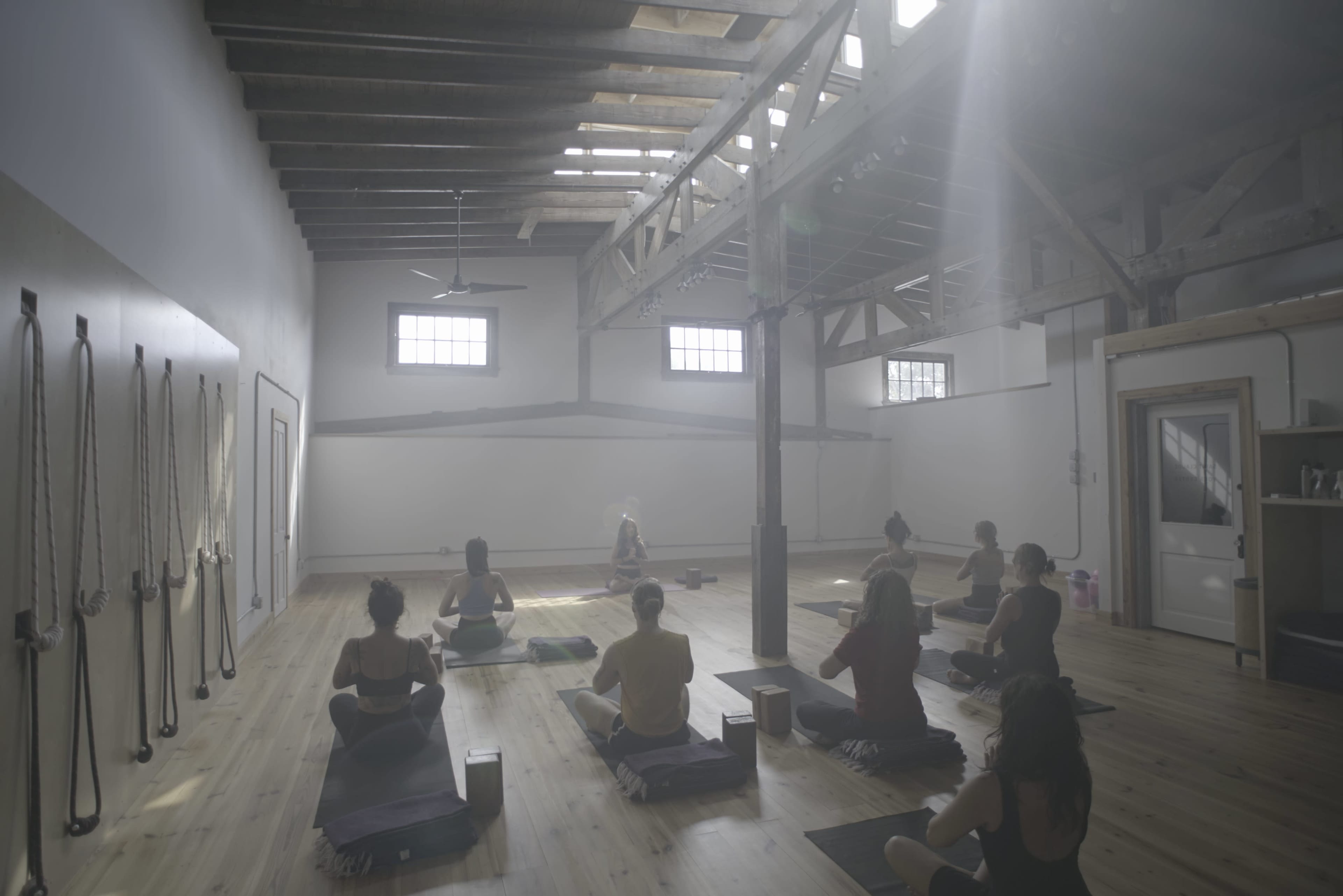 A group of people participates in a yoga class in a spacious, well-lit studio with wooden floors and beams.