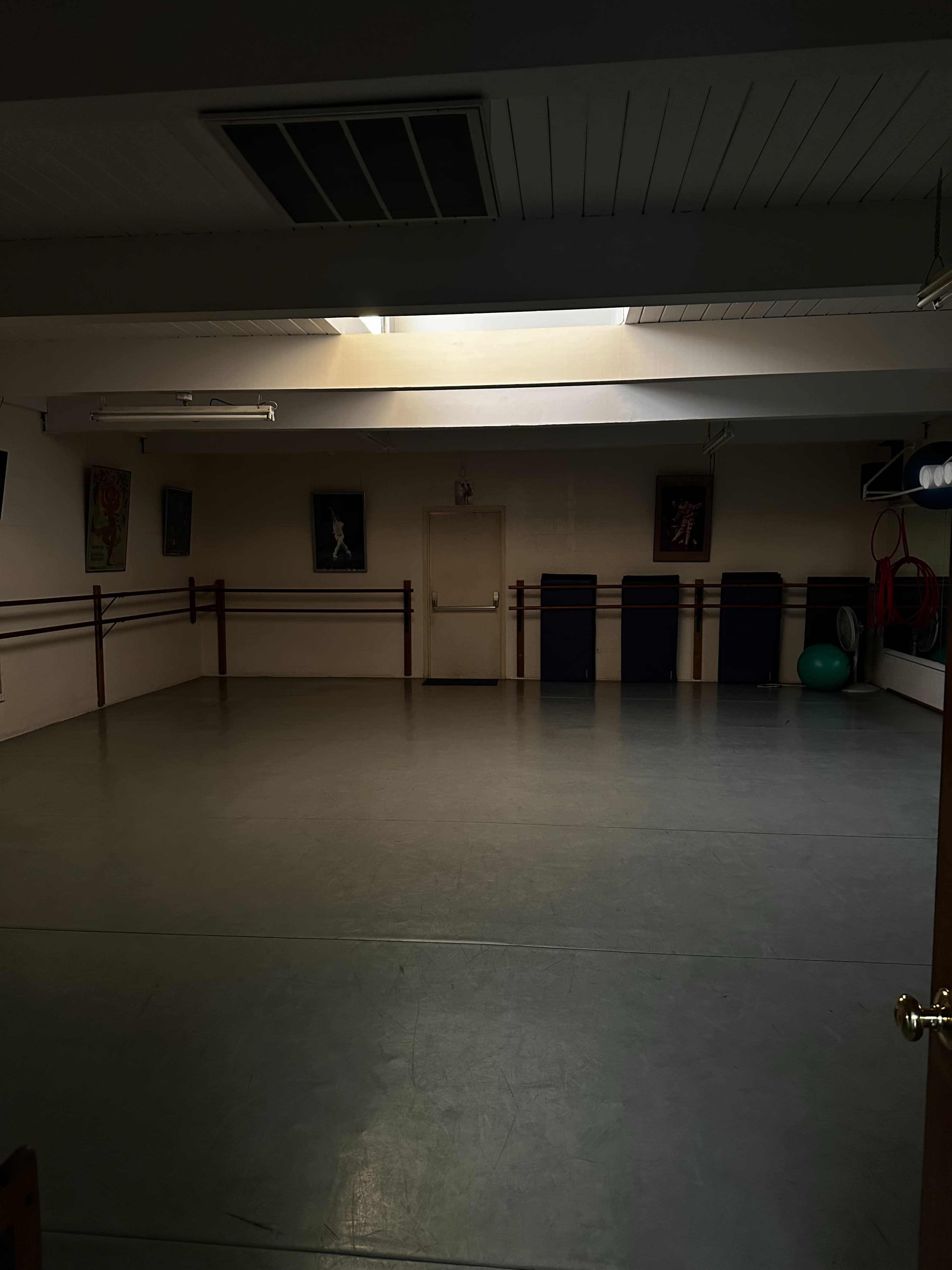The image shows an empty dance studio with a wooden floor, mirrored walls, and several exercise equipment pieces in the corners.