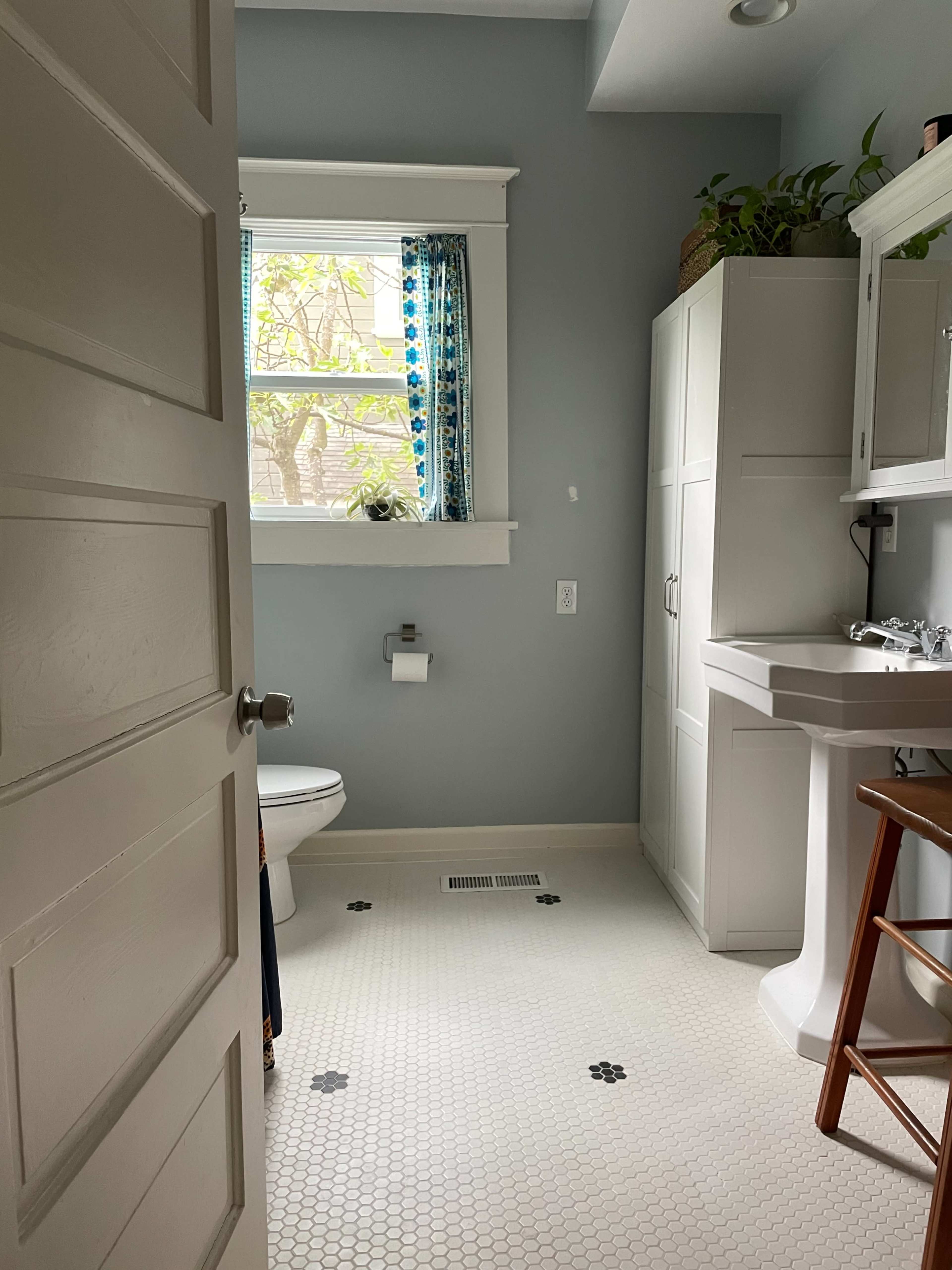 A bathroom is seen through a partially open door, featuring a toilet, sink, and a window with blue patterned curtains.
