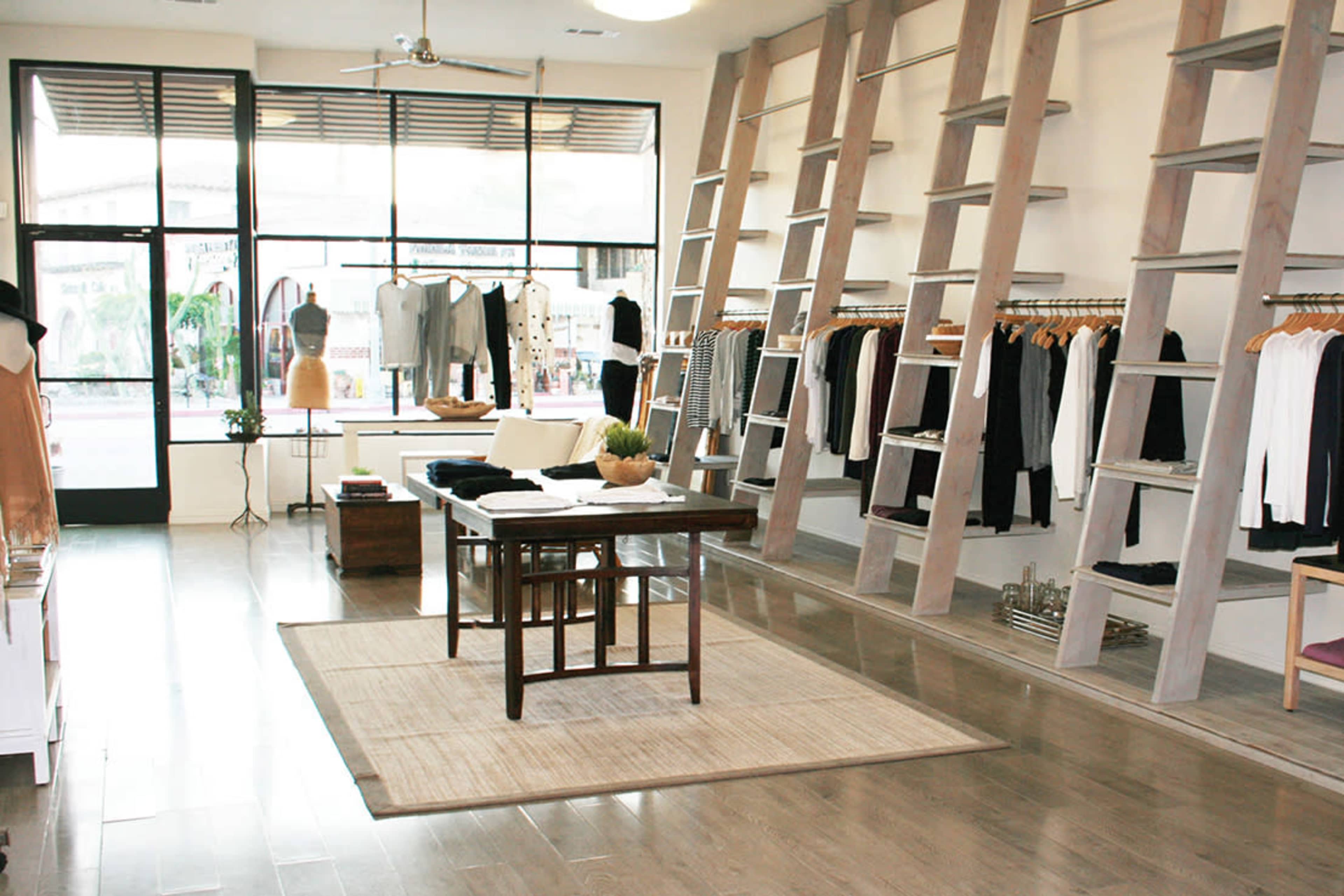 A clothing retail store featuring wooden shelving, a central table, and various garments displayed on hangers against white walls.