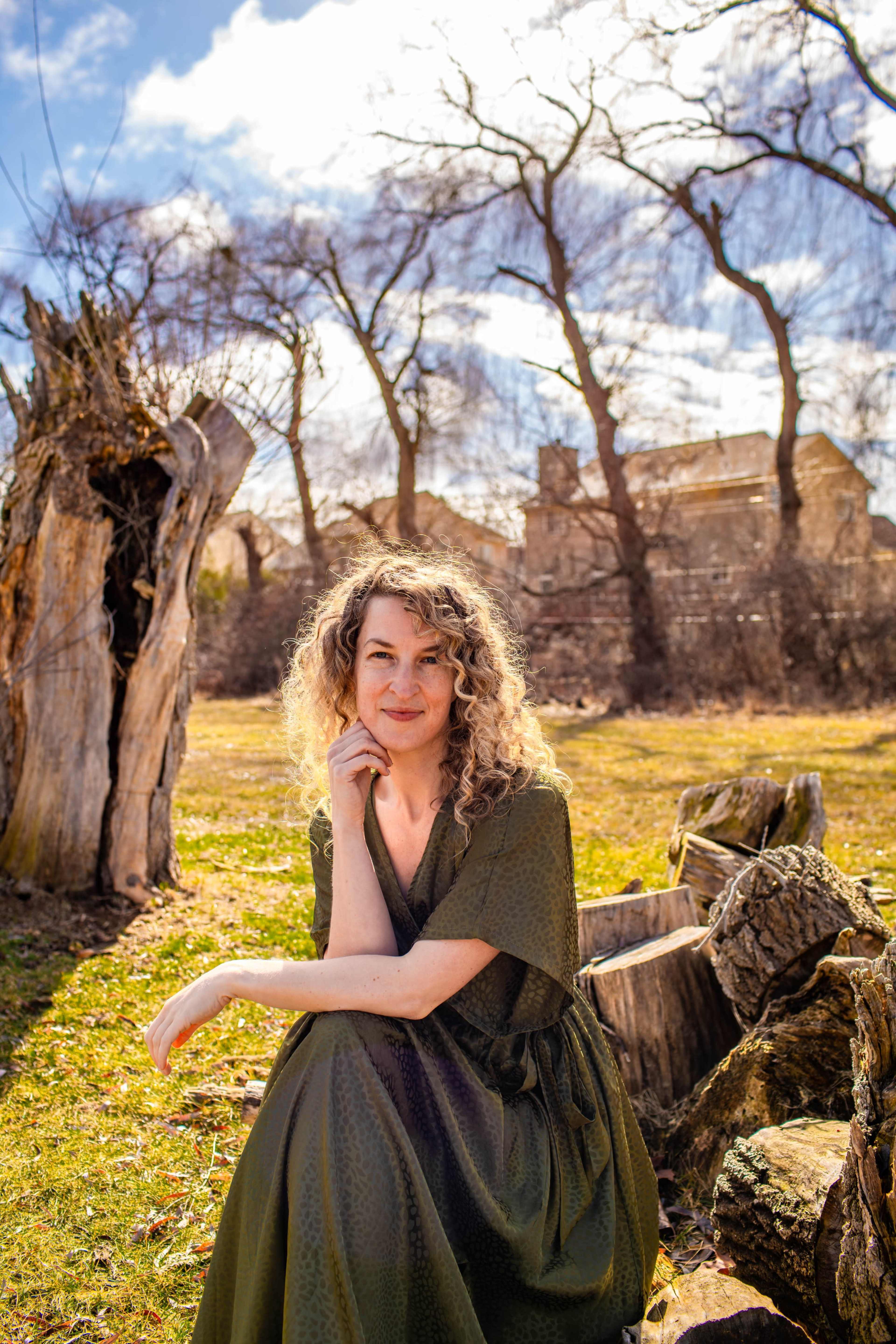 A woman with curly hair poses while sitting on a log in a grassy area with trees and a building in the background.