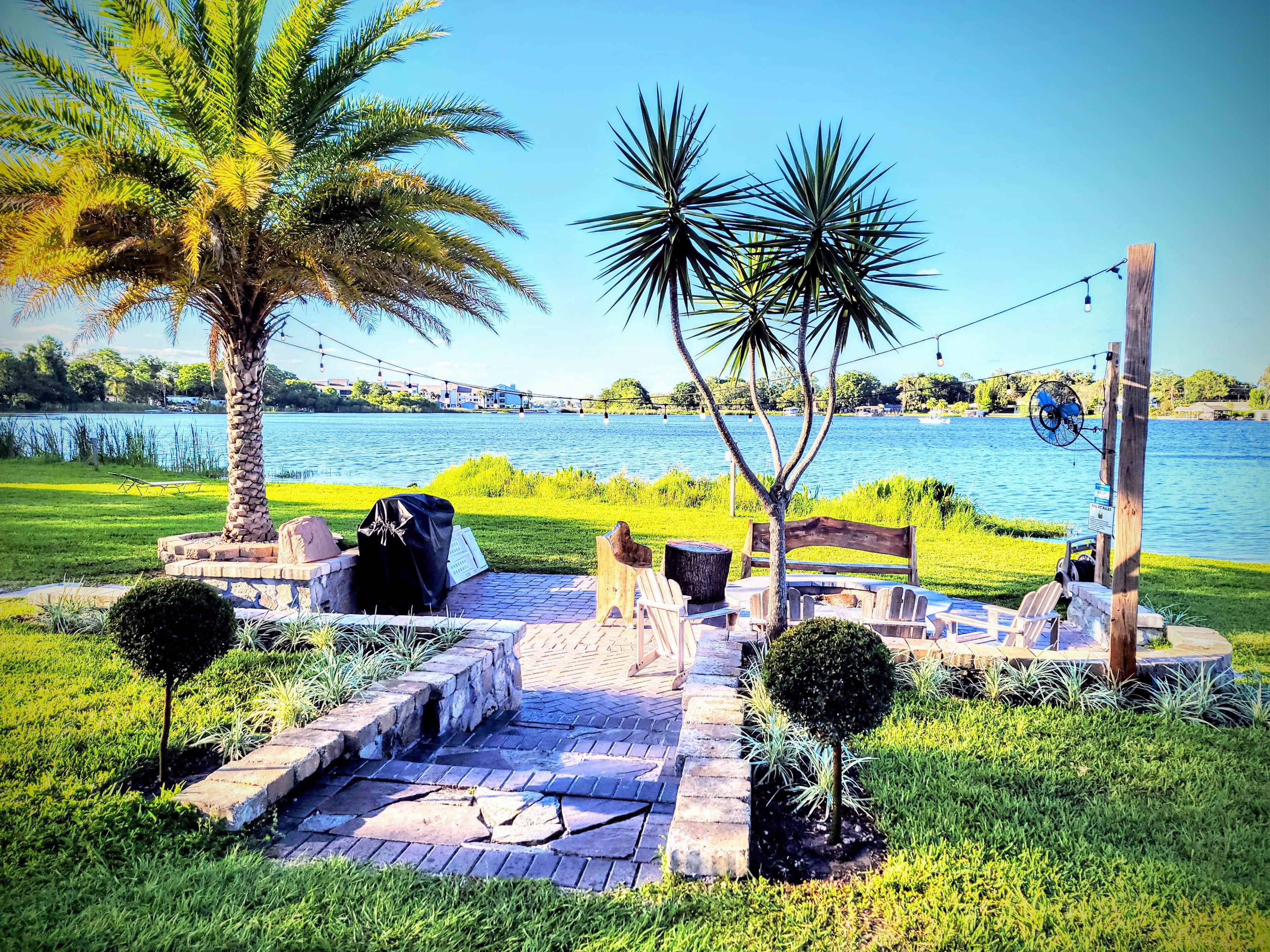 A landscaped yard by a lake, featuring a seating area with chairs and a grill, surrounded by palm trees and decorative plants.
