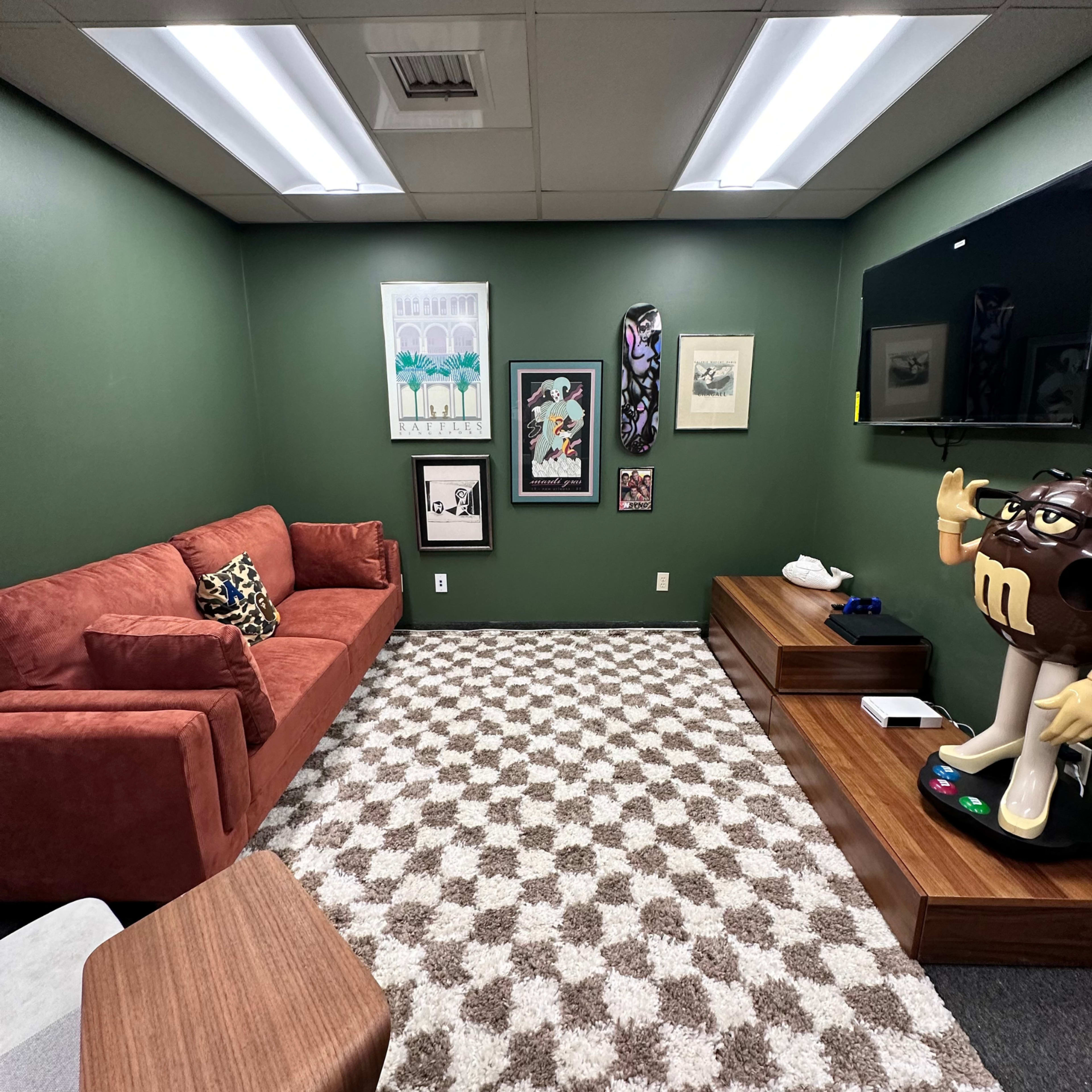 The image shows a small green-walled lounge area with a red sofa, a patterned rug, artwork on the walls, and a television mounted near a wooden furniture unit.